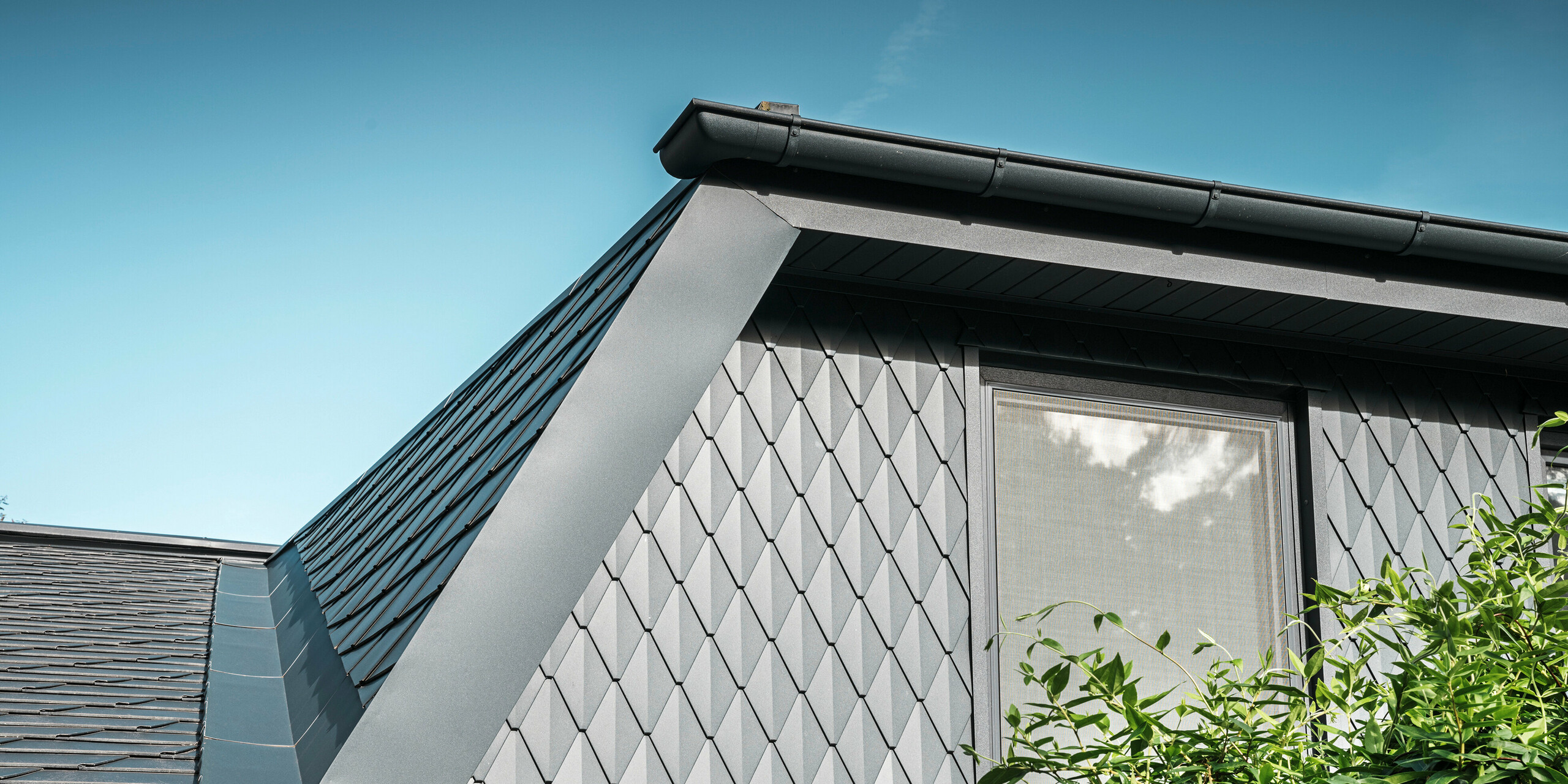 Trapezowa lukarna domu jednorodzinnego w Mechernich, pokryta małymi dachówkami PREFA w kolorze P.10 antracytowym. Dach pokryty jest gontami PREFA DS.19 w tym samym kolorze. Wysokiej jakości aluminiowa okładzina zapewnia odporność na warunki atmosferyczne oraz nowoczesny, estetyczny wygląd.