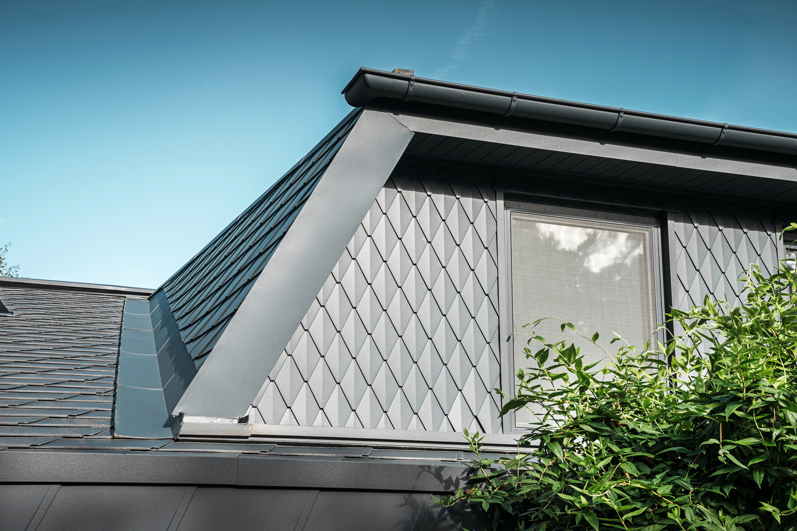 Trapezowa lukarna domu jednorodzinnego w Mechernich, pokryta małymi dachówkami PREFA w kolorze P.10 antracytowym. Dach pokryty jest gontami PREFA DS.19 w tym samym kolorze. Wysokiej jakości aluminiowa okładzina zapewnia odporność na warunki atmosferyczne oraz nowoczesny, estetyczny wygląd.