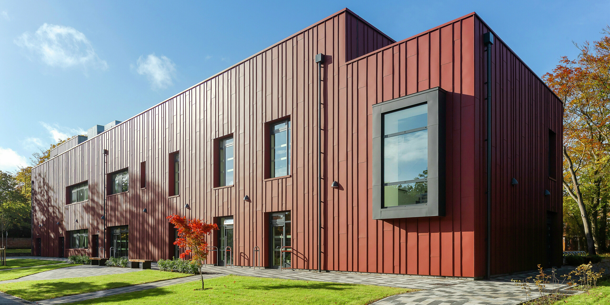 Moderne Architektur mit robuster Aluminiumfassade: Das Soldiers' Centre in Pirbright präsentiert eine elegante Stehfalzdeckung aus PREFALZ in P.10 Oxydrot, die sich harmonisch in die grüne Umgebung einfügt. Die markanten, auskragenden Fensterrahmen in P.10 Anthrazit setzen architektonische Akzente. Der gepflasterte Weg und die gepflegte Grünanlage unterstreichen die einladende Gestaltung des Gebäudes. Nachhaltiges und ästhetisches Bauen mit PREFA Aluminiumprodukten.