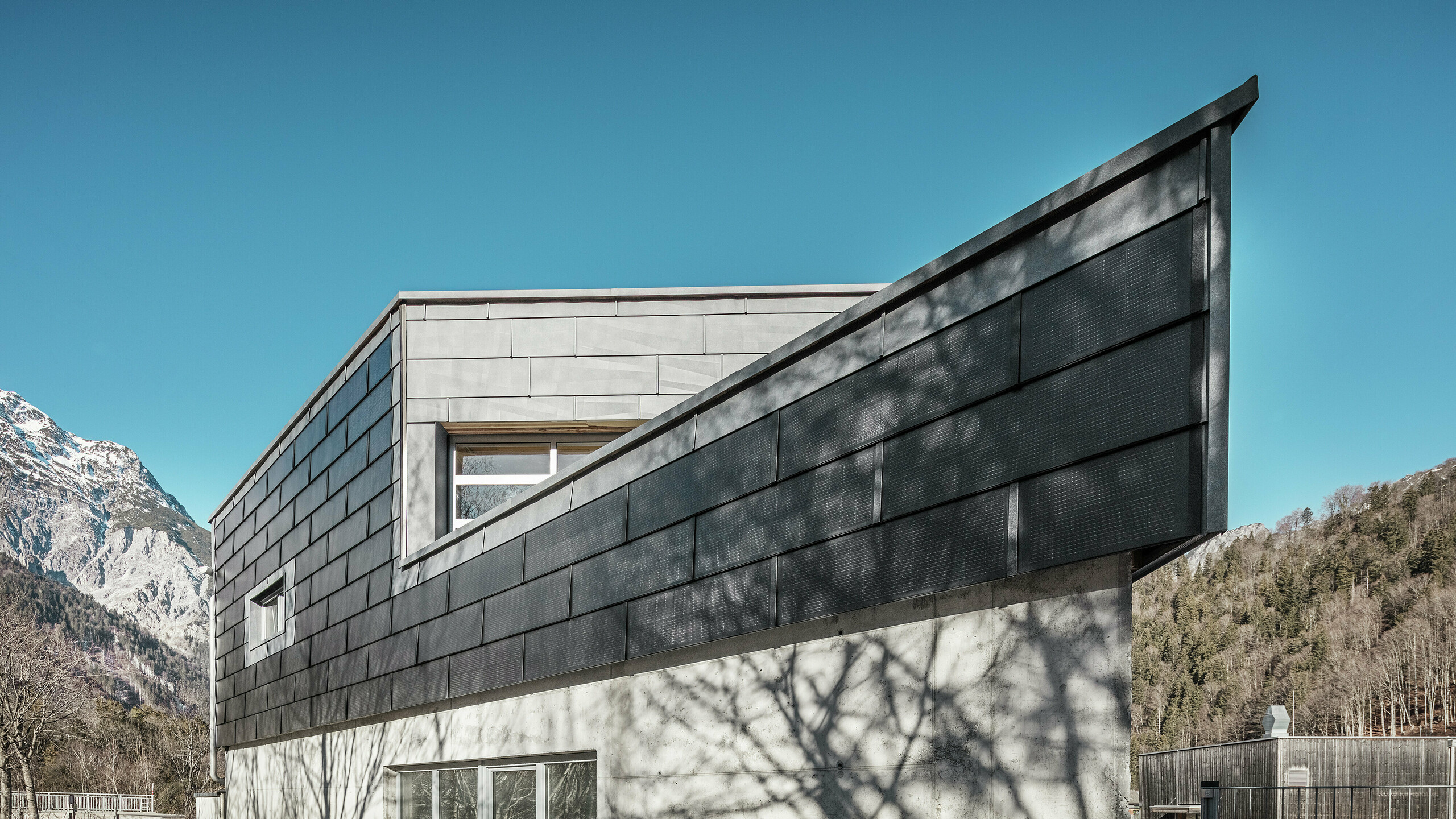 Das Bürogebäude der RÜMA Blechwerkstatt in Vorarlberg besticht durch seine moderne Fassade aus PREFA Solardachpaneelen und Sichtbeton. Umgeben von Bergen und Bäumen, deren Schatten sich an der Wand abzeichnen, erhebt es sich unter strahlend blauem Himmel. Die markante Gebäudekante und die PREFA Fassadenpaneele FX.12 unterstreichen den eindrucksvollen Eindruck zusätzlich.