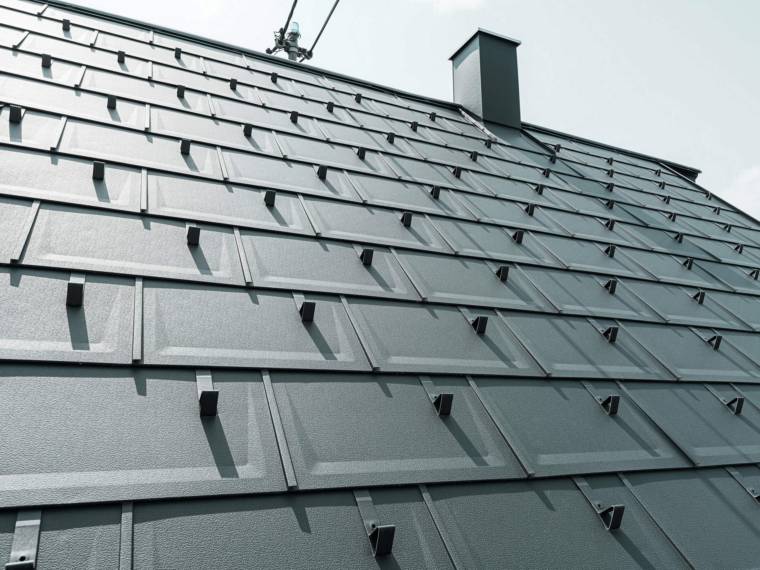 Szczegółowe zdjęcie płyty dachowej PREFA R.16 w kolorze P.10 antracyt. Na dachu widoczne są ograniczniki śniegu PREFA oraz komin.