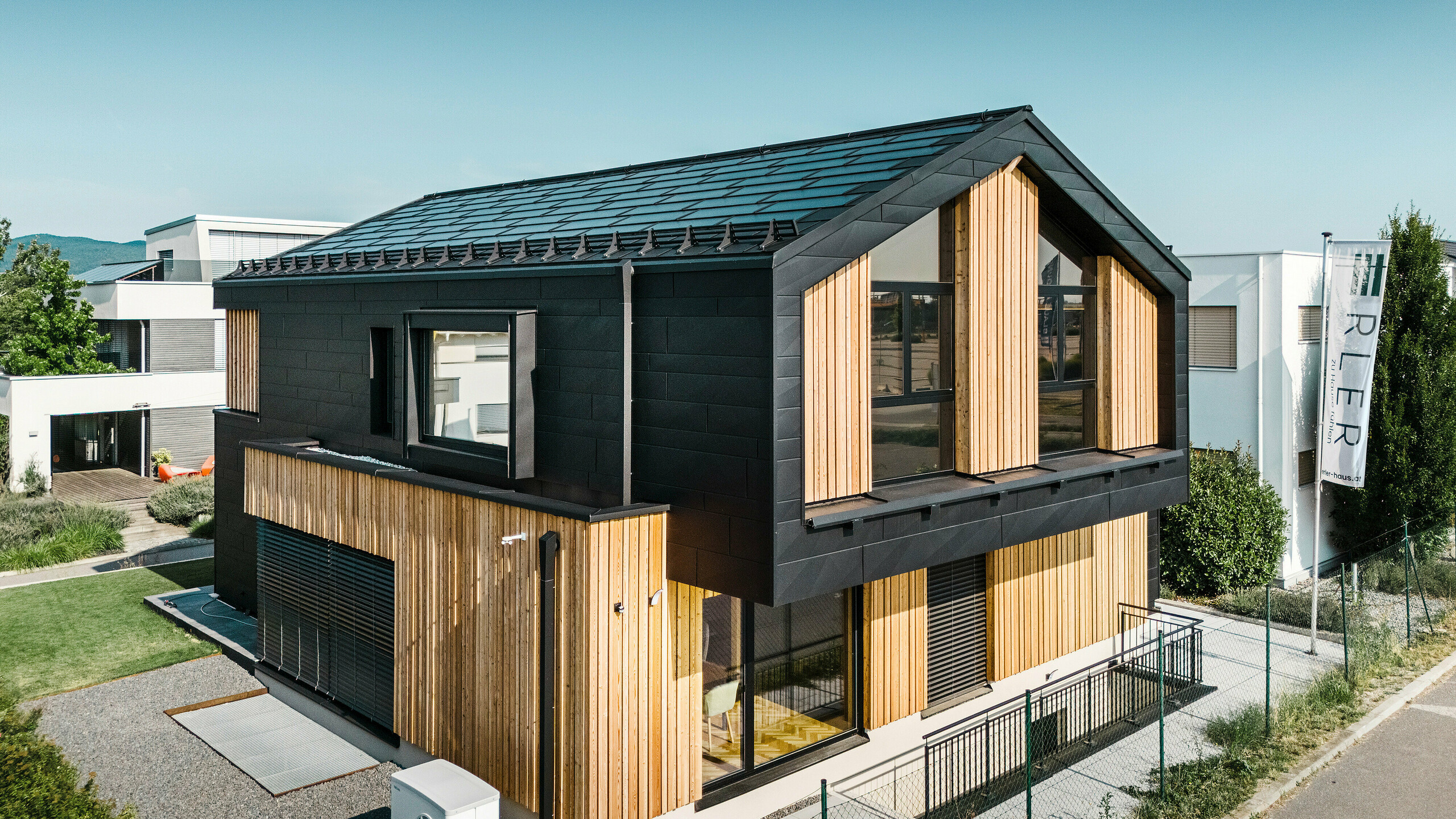 Przód domu pokazowego Autark w centrum domów prefabrykowanych Blaue Lagune, Vösendorf (AT). Dach pokryty jest czarnymi dachówkami solarnymi PREFA i dachówkami R.16 z wysokiej jakości powłoką P.10. Elewacja składa się z połączenia pionowych elementów drewnianych i PREFA Siding.X w P.10 czarnym, co nadaje budynkowi nowoczesny i ekologiczny charakter. Na prawym brzegu zdjęcia powiewa flaga firmy Erler Bau GmbH.