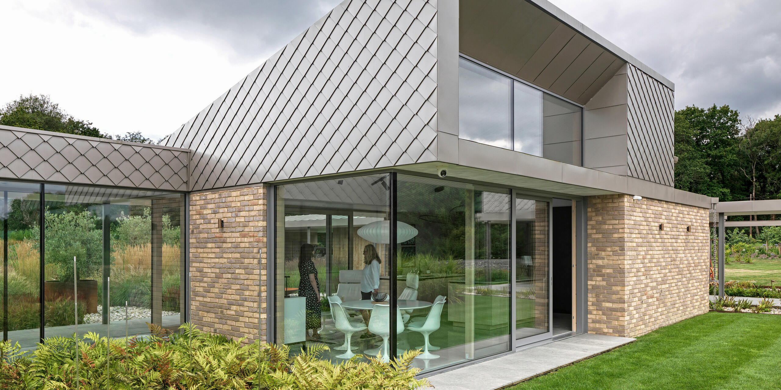 Architektonisch markanter Gebäudeteil eines Wohnhauses in West Sussex mit moderner Fassadengestaltung aus PREFA Wandrauten 20 × 20 in P.10 Bronze. Die Dachflächen sind mit PREFALZ Aluminium eingedeckt und unterstreichen durch ihre klare Linienführung den zeitlosen Charakter des Entwurfs. Großzügige Glasfronten schaffen helle Wohnräume und verbinden das Gebäude optisch mit dem gepflegten Garten. Die hochwertigen PREFA Produkte bieten Langlebigkeit und ein einheitliches Erscheinungsbild bei minimalem Wartungsaufwand.