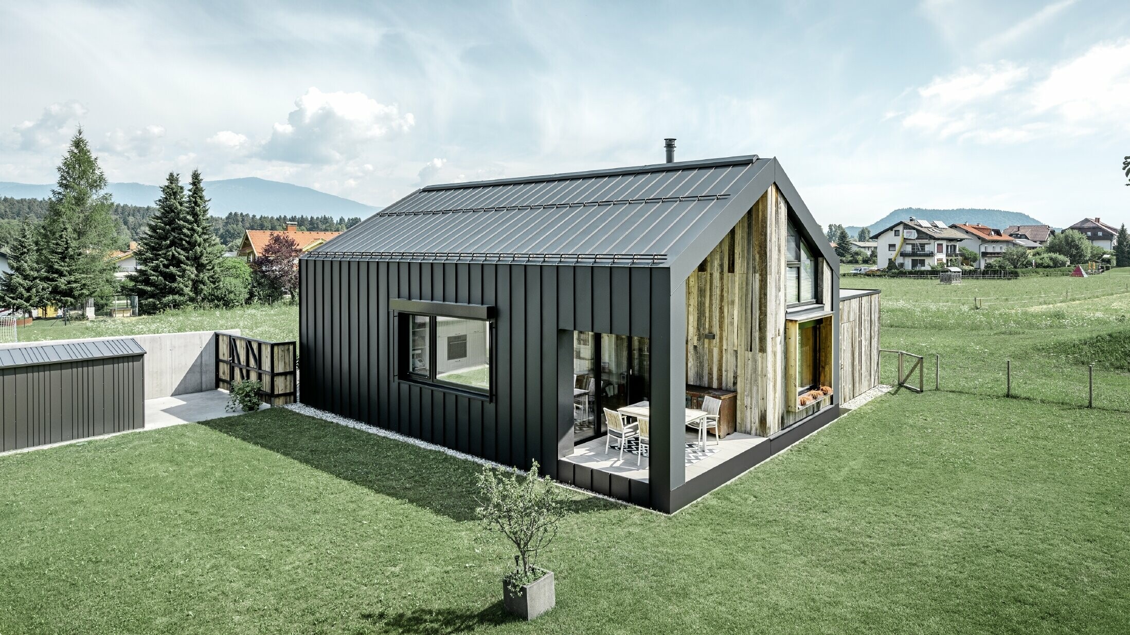 Dom jednorodzinny z dachem dwuspadowym, dach i elewacja pokryte PREFALZ od PREFA w kolorze P.10 antracytowym. Pasma przechodzą z połaci dachowej w elewację. Część elewacji pokryta jest postarzałym drewnem.