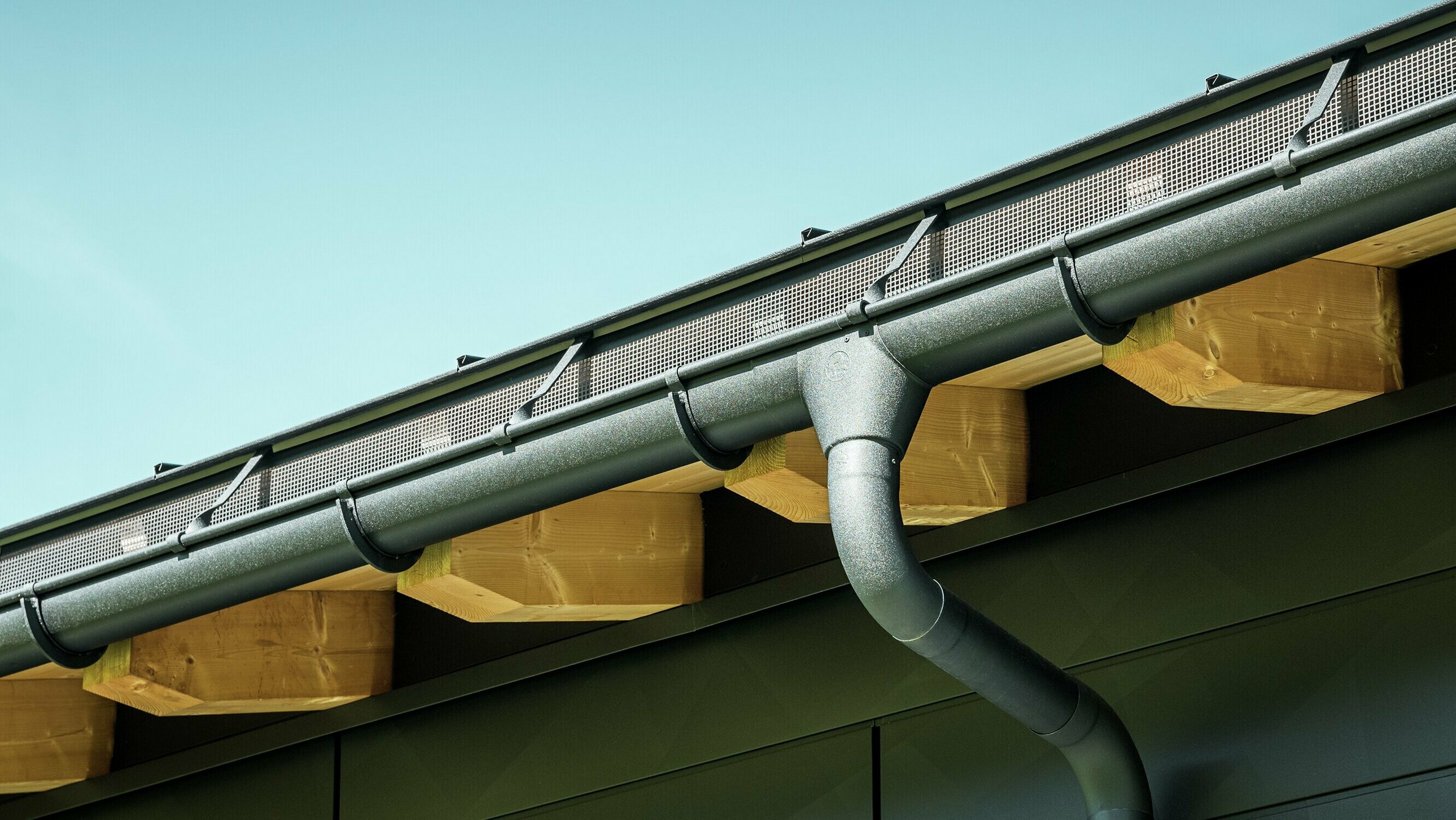 Zbliżenie elewacji nowoczesnego domu jednorodzinnego w Ascharina w Szwajcarii. Boki budynku zostały pokryte charakterystycznymi panelami elewacyjnymi PREFA Siding.X w kolorze P.10 czarny. Kolorystycznie dopasowane jest odwodnienie dachu PREFA z rynną półokrągłą i rurą spustową.