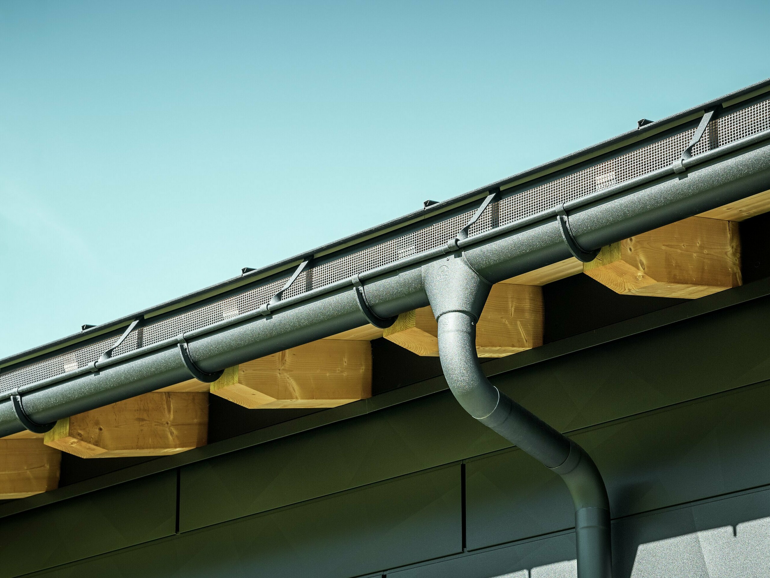 Zbliżenie elewacji nowoczesnego domu jednorodzinnego w Ascharina w Szwajcarii. Boki budynku zostały pokryte charakterystycznymi panelami elewacyjnymi PREFA Siding.X w kolorze P.10 czarny. Kolorystycznie dopasowane jest odwodnienie dachu PREFA z rynną półokrągłą i rurą spustową.