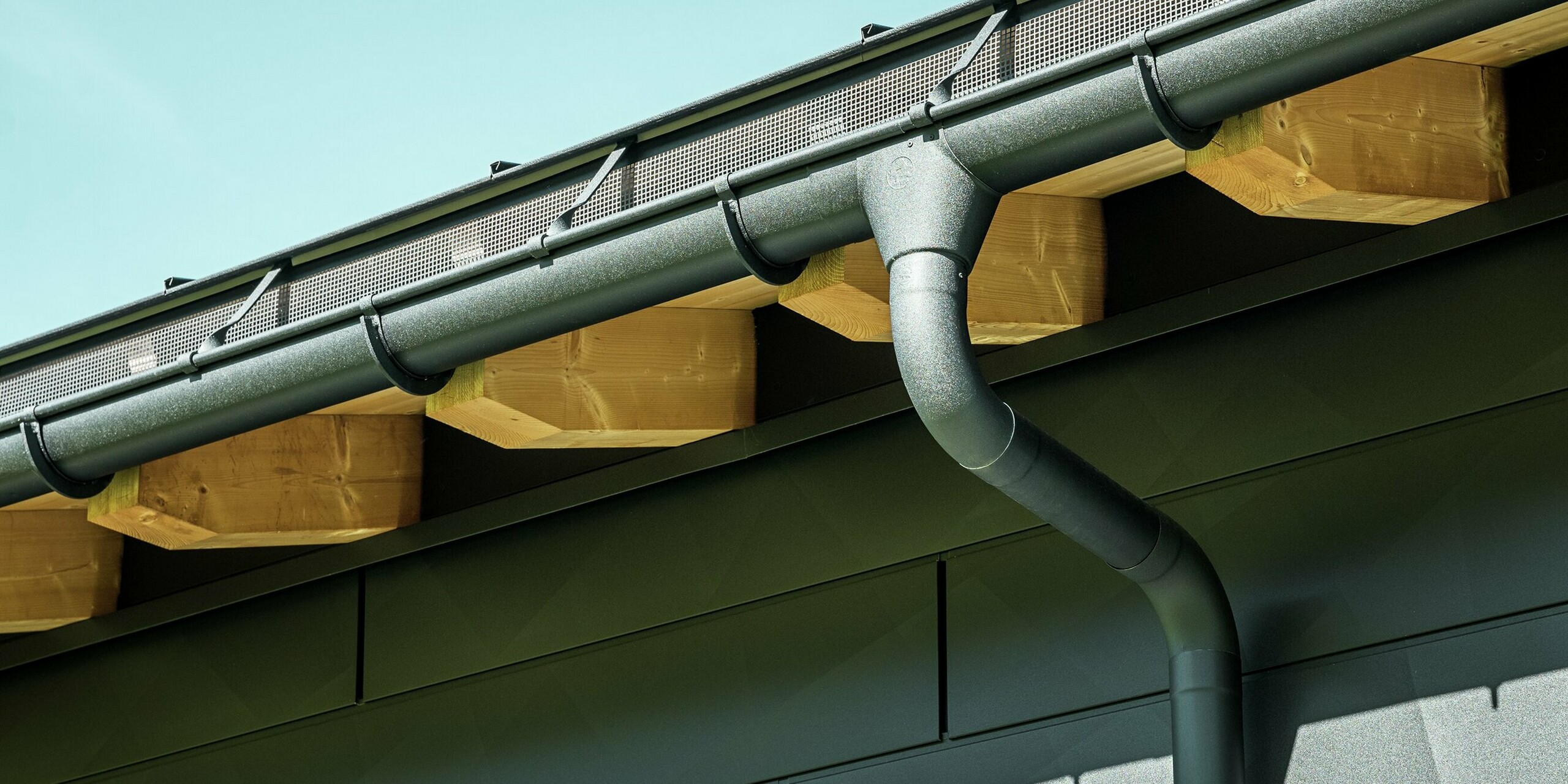 Zbliżenie elewacji nowoczesnego domu jednorodzinnego w Ascharina w Szwajcarii. Boki budynku zostały pokryte charakterystycznymi panelami elewacyjnymi PREFA Siding.X w kolorze P.10 czarny. Kolorystycznie dopasowane jest odwodnienie dachu PREFA z rynną półokrągłą i rurą spustową.