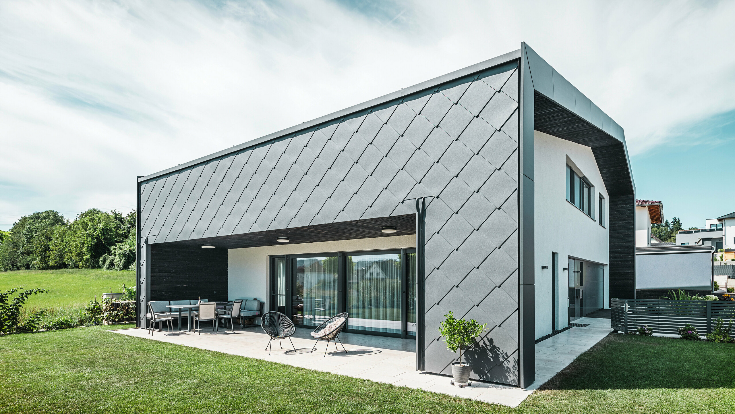 Zadbany ogród przy domu jednorodzinnym w Aschach. Budynek jest stylowo pokryty grafitowymi rombami elewacyjnymi PREFA Aluminium 44 × 44. Zadaszony taras stanowi przytulne miejsce do odpoczynku z widokiem na soczystą zieloną trawnik. Elegancka okładzina aluminiowa w połączeniu z białymi tynkami zapewnia nowoczesną i odporną na warunki atmosferyczne architekturę. Duże powierzchnie okienne wpuszczają do wnętrza dużo światła dziennego i tworzą otwartą, jasną atmosferę.