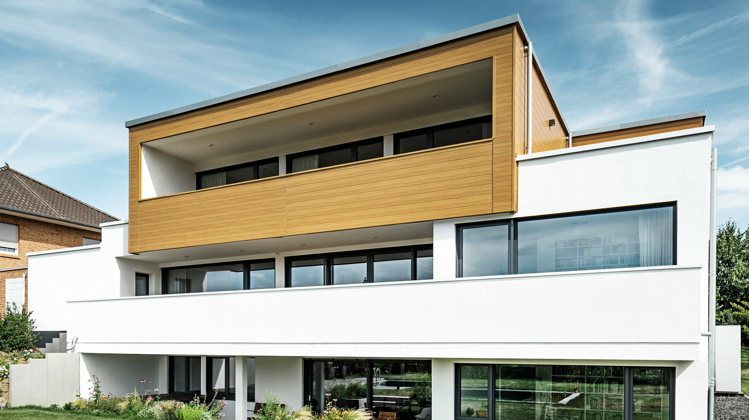 Tył nowoczesnego domu jednorodzinnego w Usingen w Niemczech. W dolnej części zdjęcia widać zielony trawnik, basen i młode drzewo ogrodowe. Przyciągający wzrok dom ma białą podstawę budynku wykonaną z otynkowanej fasady, aluminiową fasadę na piętrze wykonaną z PREFA Sidings w kolorze dąb naturalny i duże powierzchnie okienne z ramą w kolorze antracytu. Sąsiedni dom z ceglaną fasadą jest widoczny z daleka po lewej stronie zdjęcia.