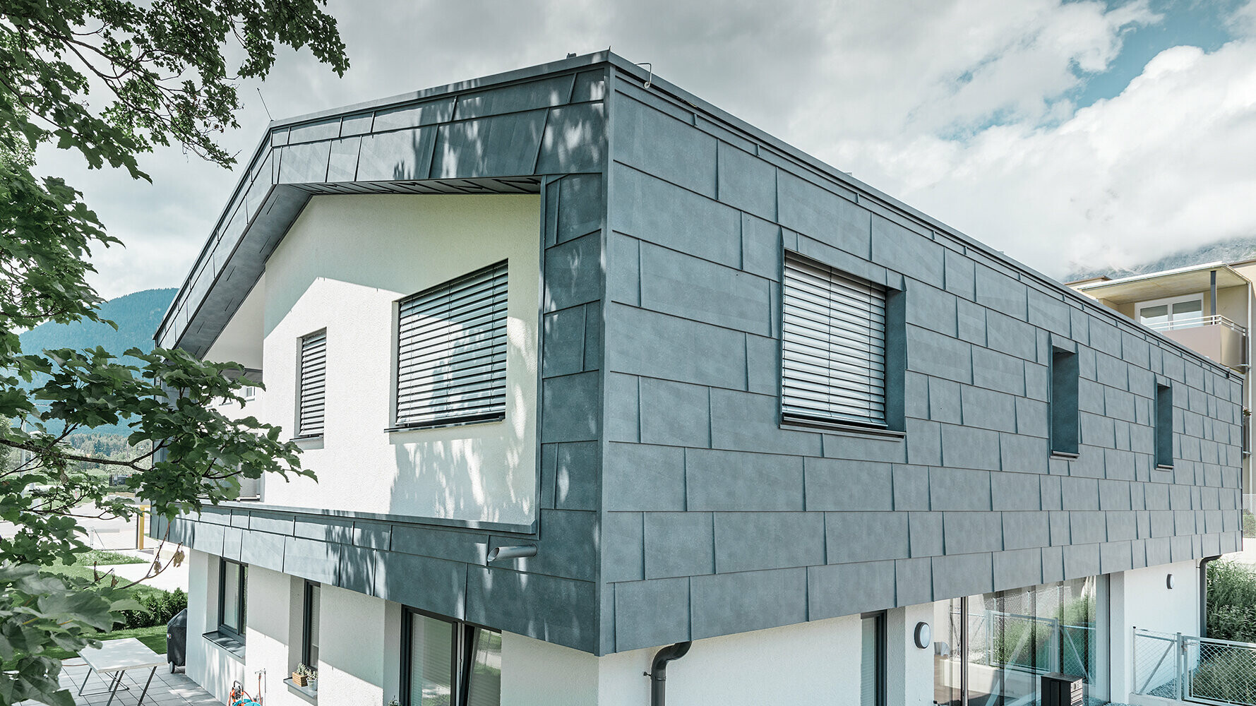 Nowoczesny dom jednorodzinny bez okapu dachowego; górna kondygnacja pokryta panelami elewacyjnymi PREFA FX.12 w kolorze szarym kamiennym; pozostała część elewacji otynkowana na biało.