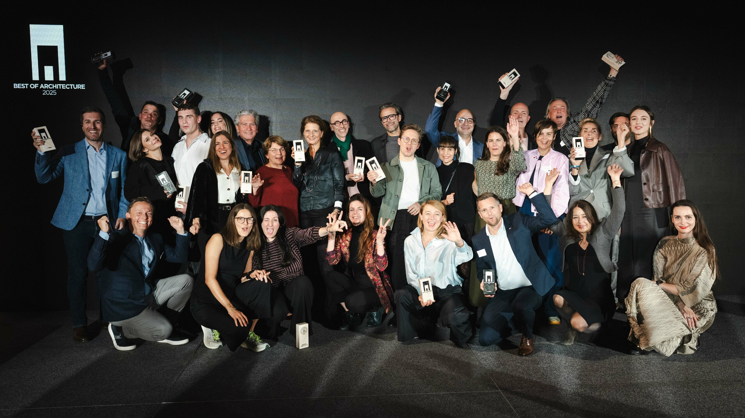 Zdjęcie grupowe zwycięzców podczas ceremonii wręczenia nagród „Budynek mieszkalny roku 2025”: zwycięzcy stoją przed elegancką czarną sceną z logo „Best of Architecture” i dumnie trzymają w rękach swoje nagrody. Dynamiczne pozy, nowoczesne ubrania i efektowne oświetlenie podkreślają znaczenie wydarzenia, a scena symbolizuje innowacyjność i doskonałość architektoniczną.