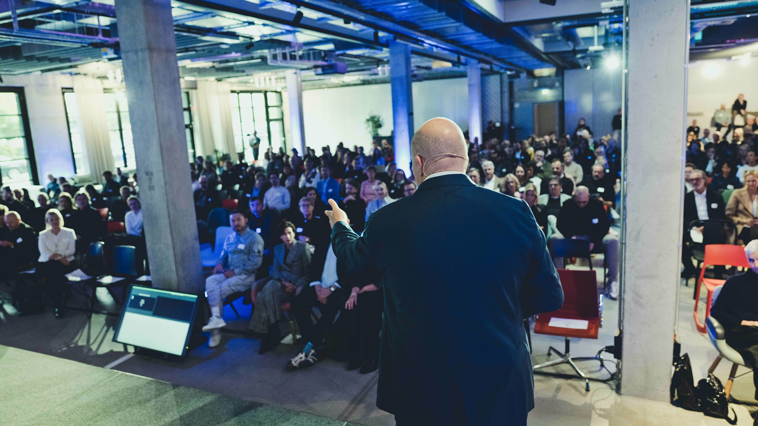 Ciekawa scena z wykładu podczas ceremonii wręczenia nagród „Budynek mieszkalny roku 2025”: widok ze sceny na wypełnioną po brzegi salę w nowoczesnym, pełnym światła pomieszczeniu o industrialnym wystroju. Na pierwszym planie wykładowca stoi przed dużym ekranem, a uważni słuchacze śledzą prezentację. Wydarzenie poświęcone jest innowacjom i aktualnym trendom w architekturze.