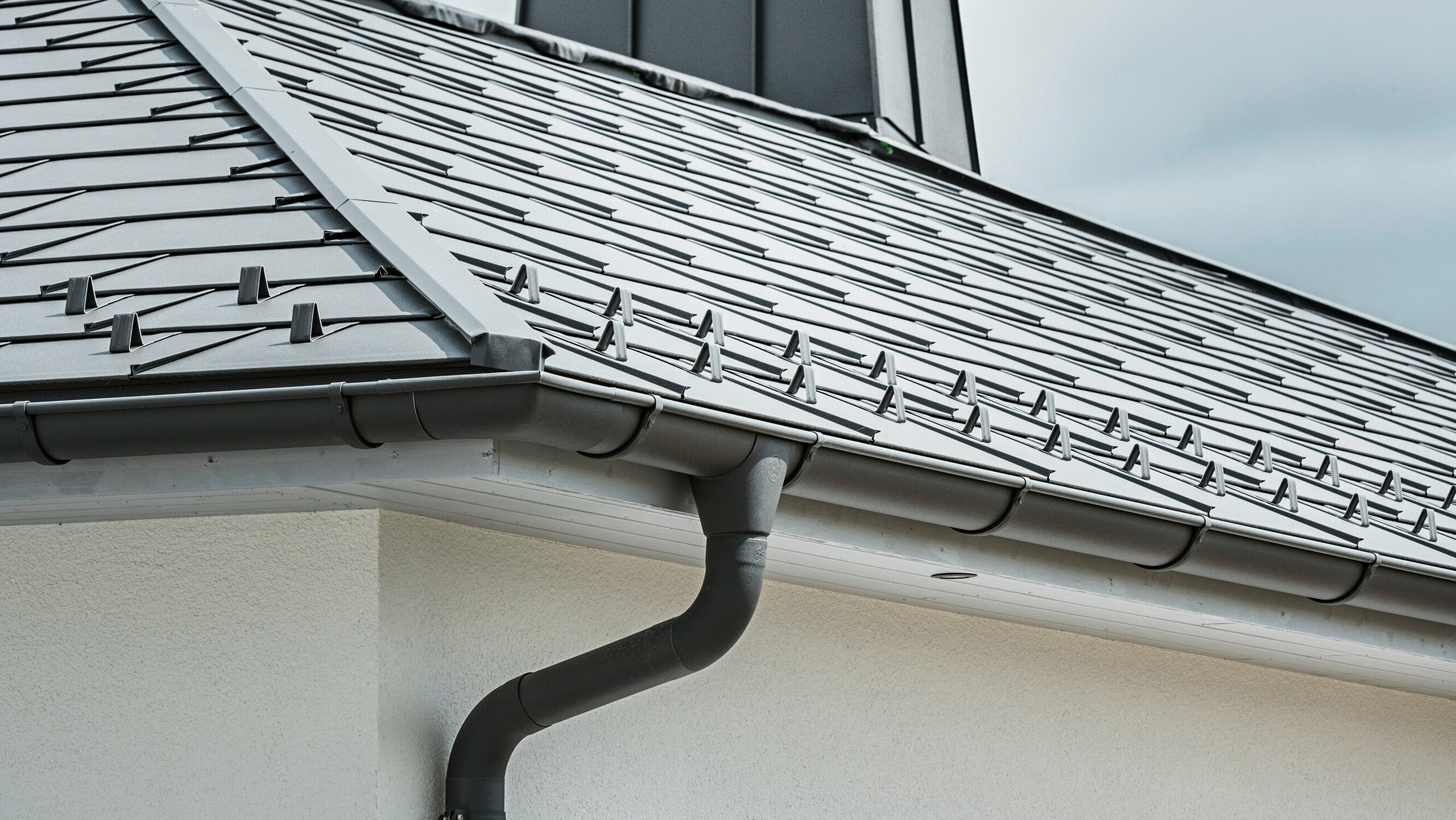 Zbliżenie na okap domu jednorodzinnego wyposażonego w wydajny system odprowadzania wody deszczowej PREFA w kolorze P.10 grafitowym. Zdjęcie przedstawia szczegółowo ułożoną dachówkę PREFA R.16, a pod nią bezszwowo zintegrowaną rynnę półokrągłą oraz rurę spustową, które razem stanowią niezawodne i estetyczne rozwiązanie do odprowadzania wody deszczowej. Wytrzymałe produkty aluminiowe i staranny montaż podkreślają jakość i trwałość kompletnego systemu PREFA.