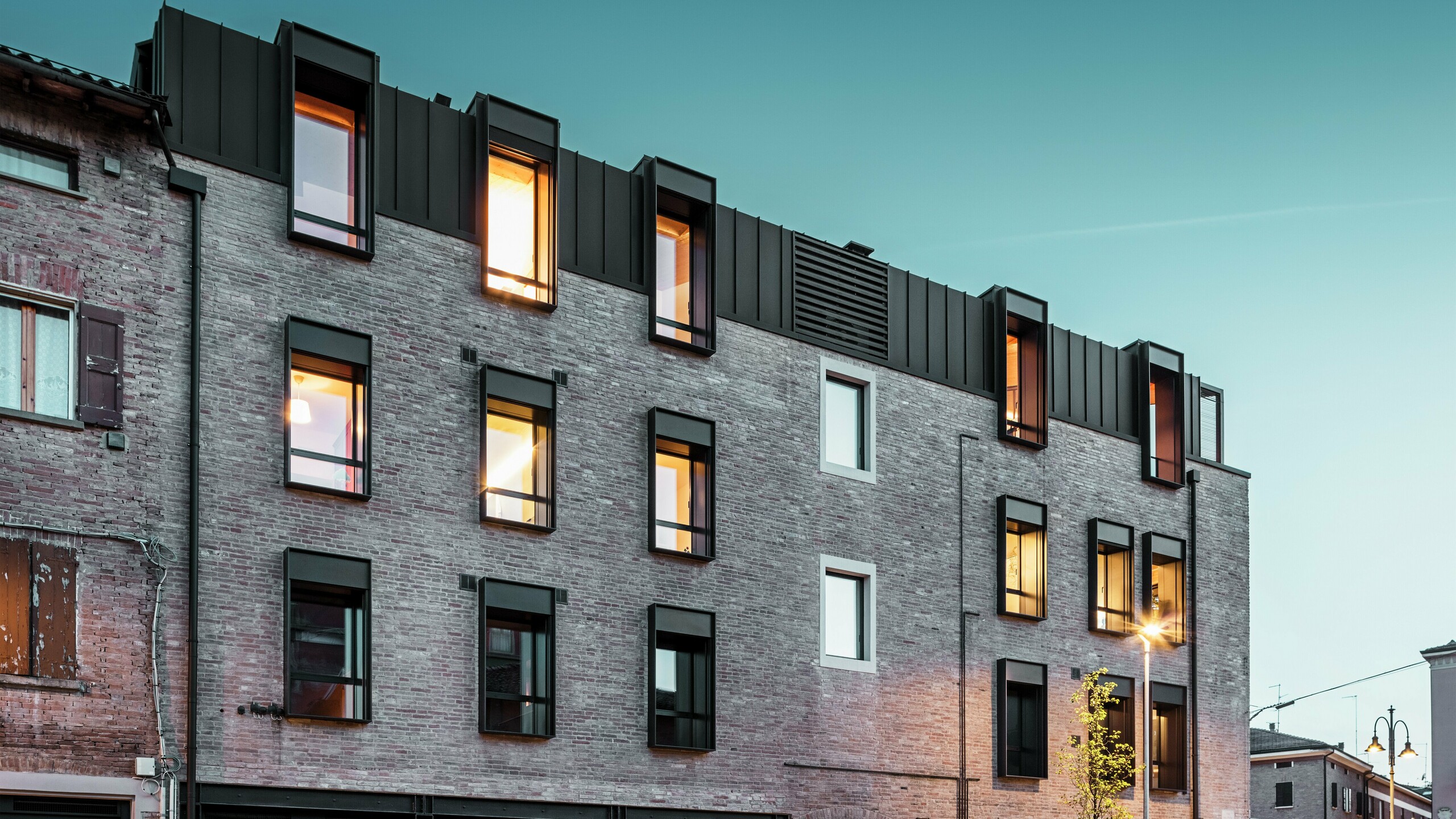 Miejski budynek z cegły we Włoszech z nowoczesnym dachem aluminiowym PREFALZ w kolorze P.10 orzechowy brąz, oświetlonymi oknami i otaczającą go historyczną architekturą o zmierzchu.