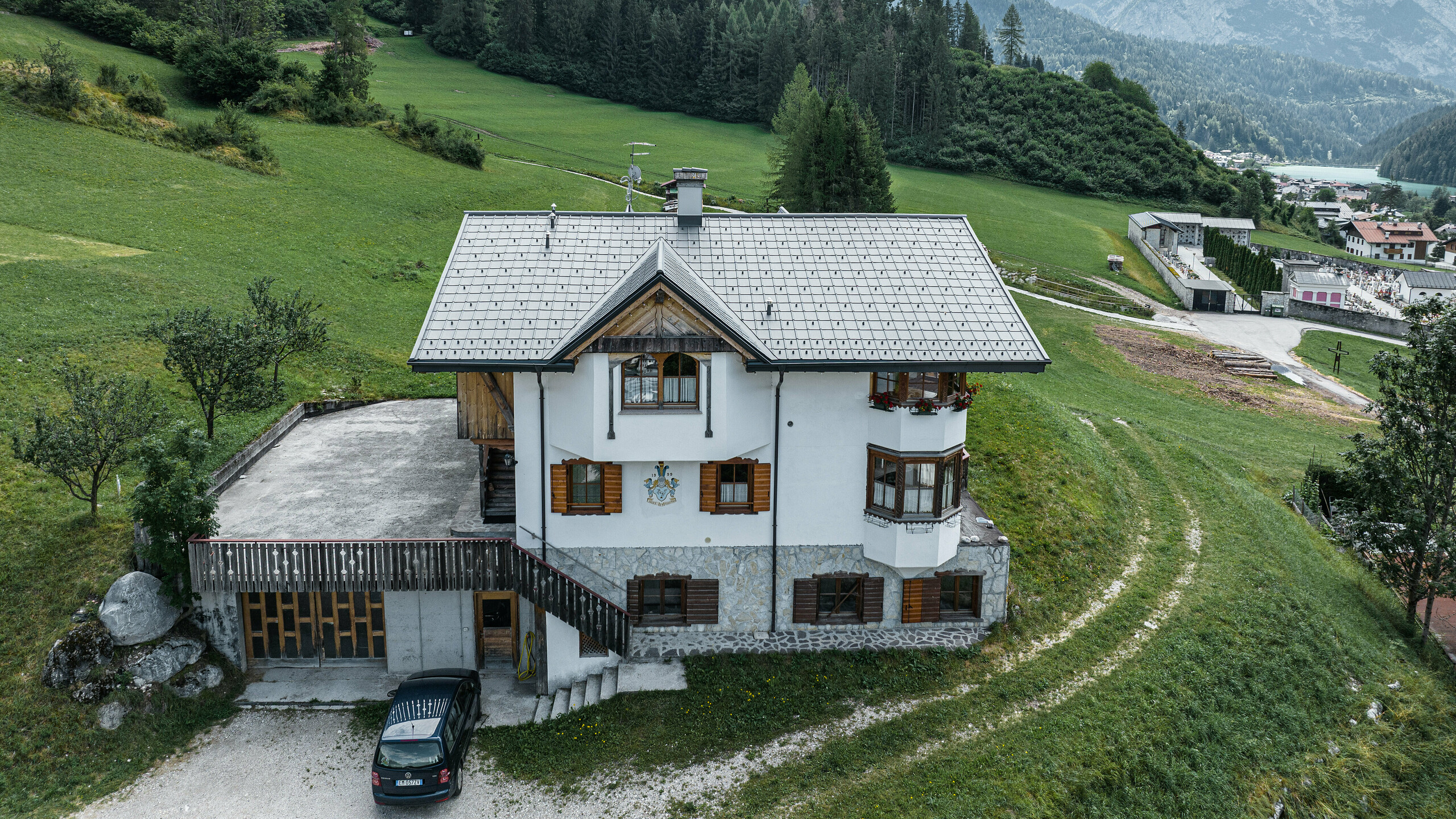 Blick aus der Vogelperspektive auf den Eingang der traditionellen Berghütte in Auronzo di Cadore nach der Dachsanierung mit PREFA Ziegeln aus Aluminium in P.10 Hellgrau. Das Gebäude mit seinem charakteristischen alpinen Charme und dem modernen Blechdach fügt sich harmonisch in die üppig grüne Landschaft und das beeindruckende Bergpanorama im Hintergrund ein und demonstriert eine gelungene Kombination aus Funktionalität und regionaler Architektur.