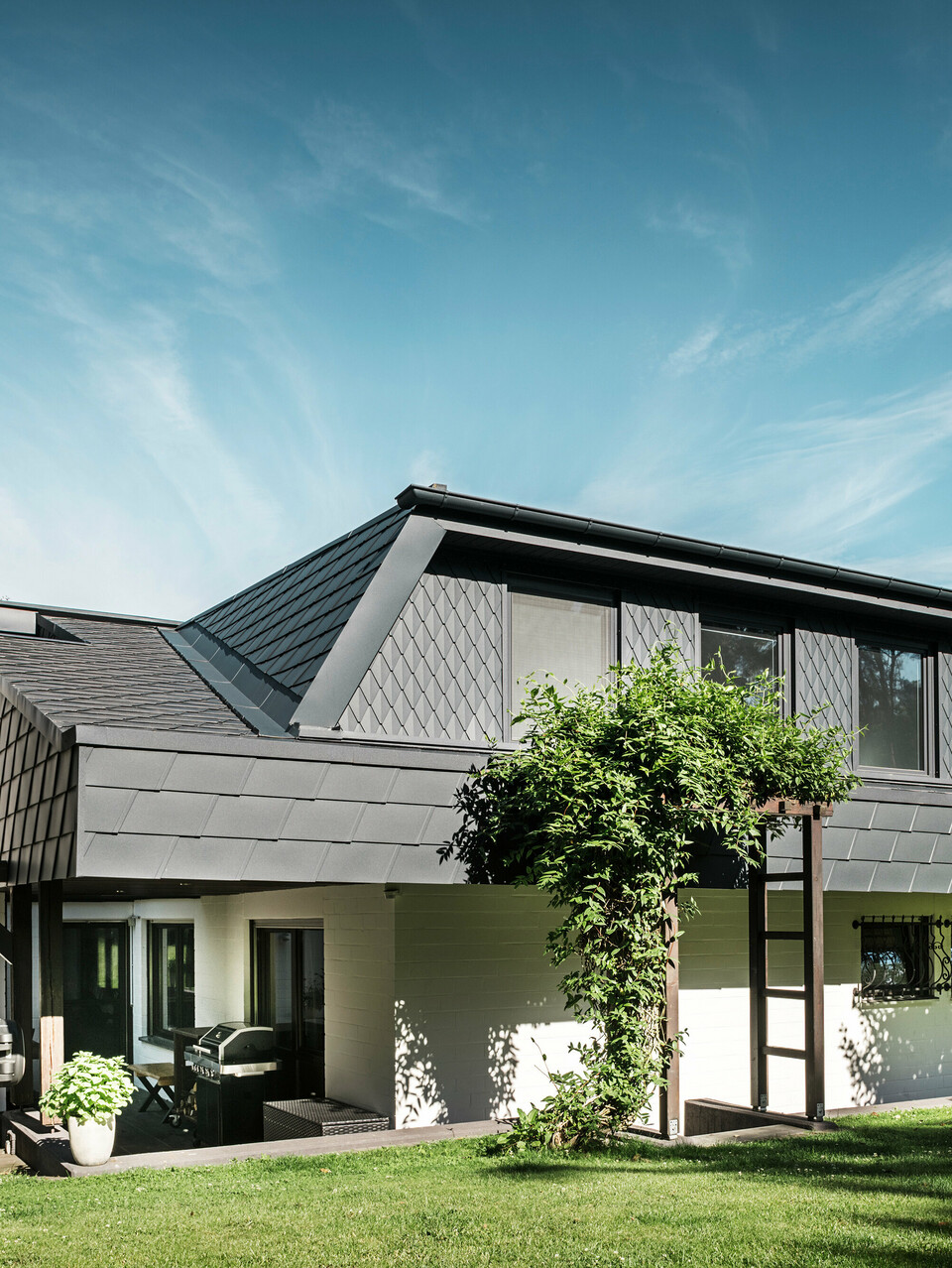 Seitliche Ansicht eines Einfamilienhauses in Mechernich mit PREFA Dachschindeln DS.19 und kleinen Dachrauten in P.10 Anthrazit. Die Trapezgauben sind mit Dachrauten aus robustem Blech verkleidet, während das Dach mit Schindeln gedeckt ist. Eine grüne Pergola und Bäume im Gartenbereich ergänzen die moderne und wetterbeständige Architektur.