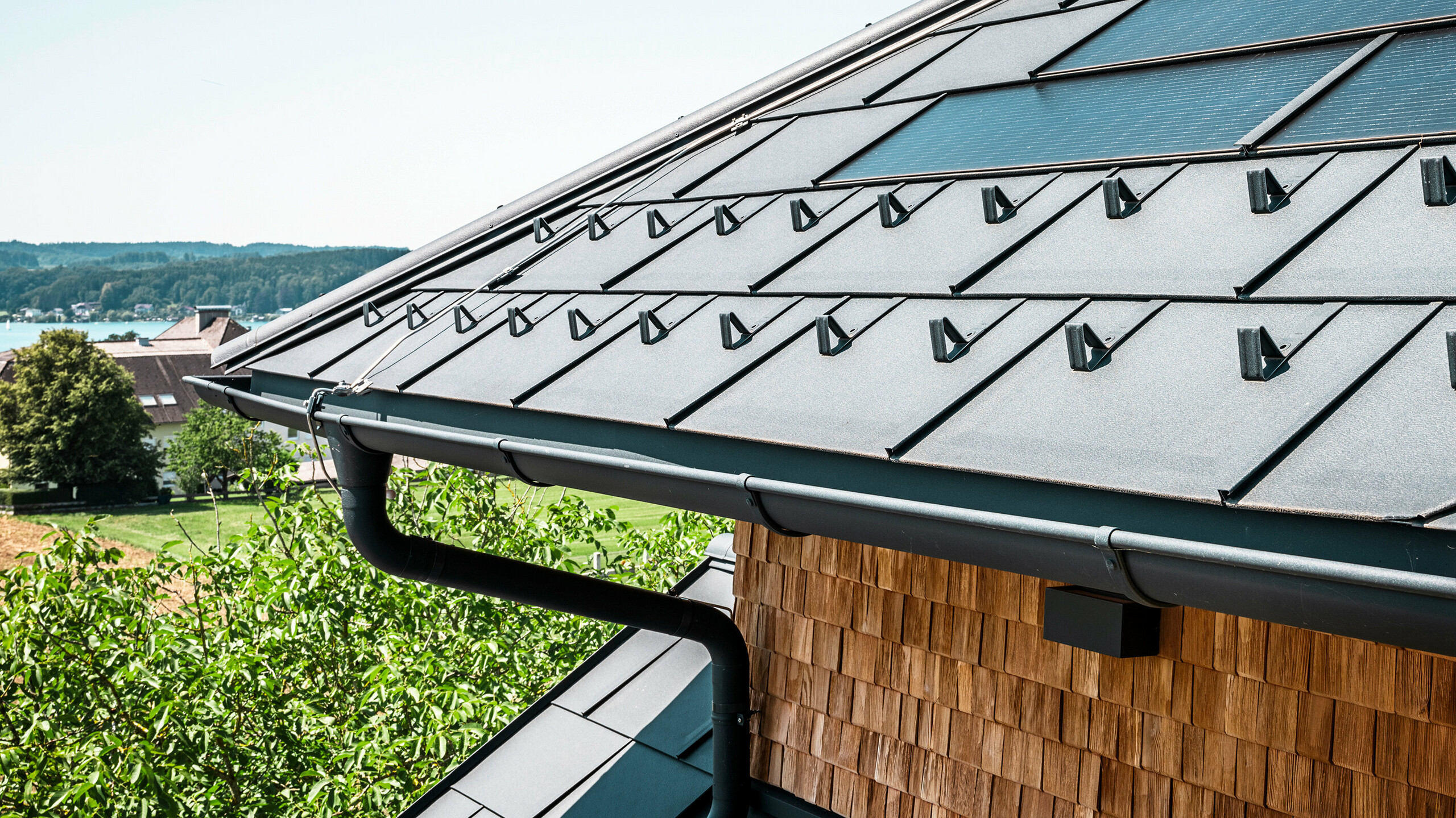 Restauracja Zenz'n Stub'n w Schörfling am Attersee prezentuje nowo wyremontowany dach z innowacyjnymi dachówkami solarnymi PREFA i trwałymi dachówkami PREFA R.16 w kolorze P.10 antracyt. Stylowy dach aluminiowy łączy w sobie nowoczesny design z zrównoważonym pozyskiwaniem energii. Również system odwodnienia dachu, składający się z rury spustowej i rynny półokrągłej w kolorze P.10 antracyt, harmonijnie komponuje się z całością i podkreśla wysoką jakość wykonania. W tle mieni się błękit jeziora Attersee.