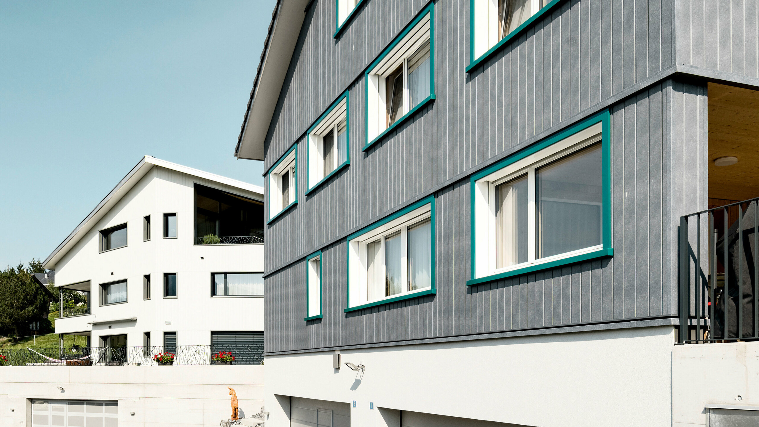 Ansicht zweier benachbarter Mehrfamilienhäuser in Weissbad, Schweiz, wobei das vordere Gebäude mit seiner PREFA Siding Fassade in P.10 Steingrau einen starken visuellen Eindruck vermittelt. Die vertikal montierten Sidings schaffen eine elegante und schützende Hülle, während die türkisen Fensterrahmen farbliche Akzente setzen. Das Bild fängt das Zusammenspiel der modernen Materialien mit der traditionellen Architektur ein und demonstriert die vielseitige Einsetzbarkeit von PREFA Aluminiumprodukten.