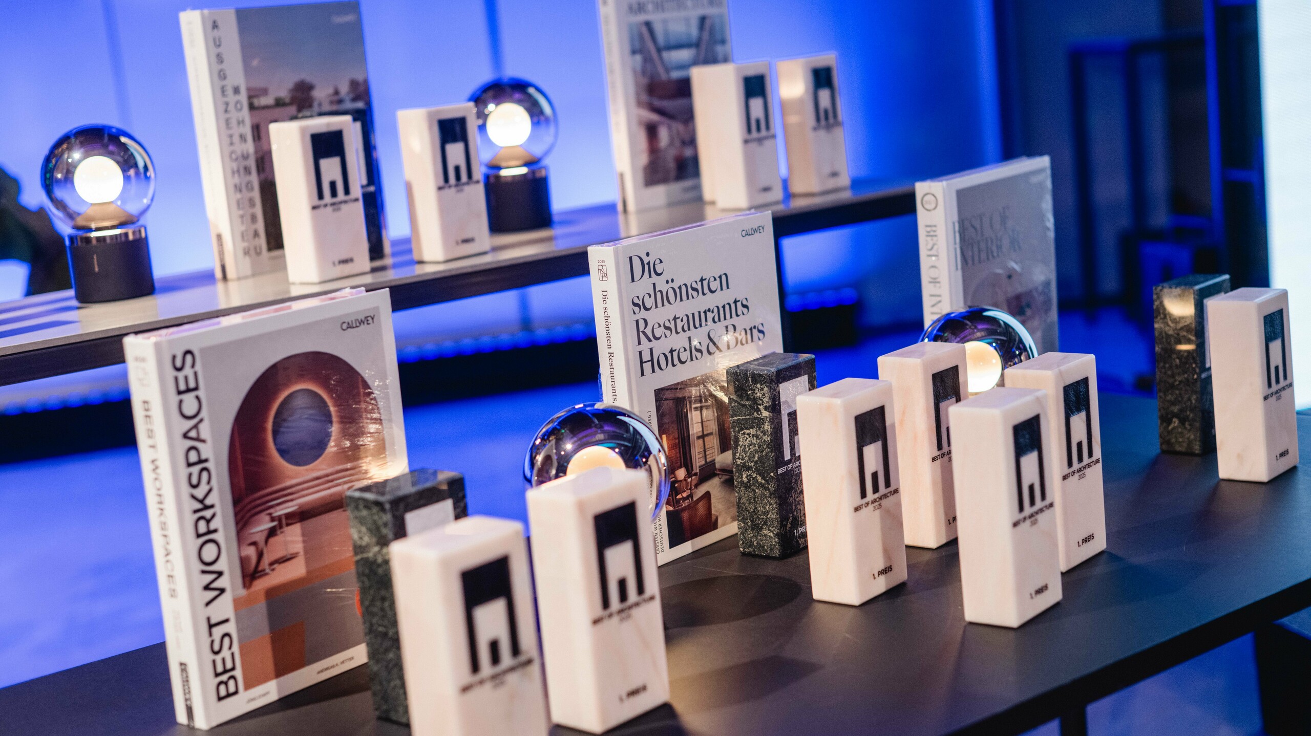 Wystawa nagród z konkursu „Budynek mieszkalny roku” 2025: kilka eleganckich trofeów z logo „Best of Architecture” jest wyeksponowanych na nowoczesnym stole, obok wysokiej jakości książek o architekturze i designie, takich jak „Best Workspaces” i „Die schönsten Restaurants Hotels & Bars” (Najpiękniejsze restauracje, hotele i bary). Scena jest skąpana w nastrojowym niebieskim świetle, które oddaje ekskluzywność, innowacyjność i doskonałość architektoniczną.