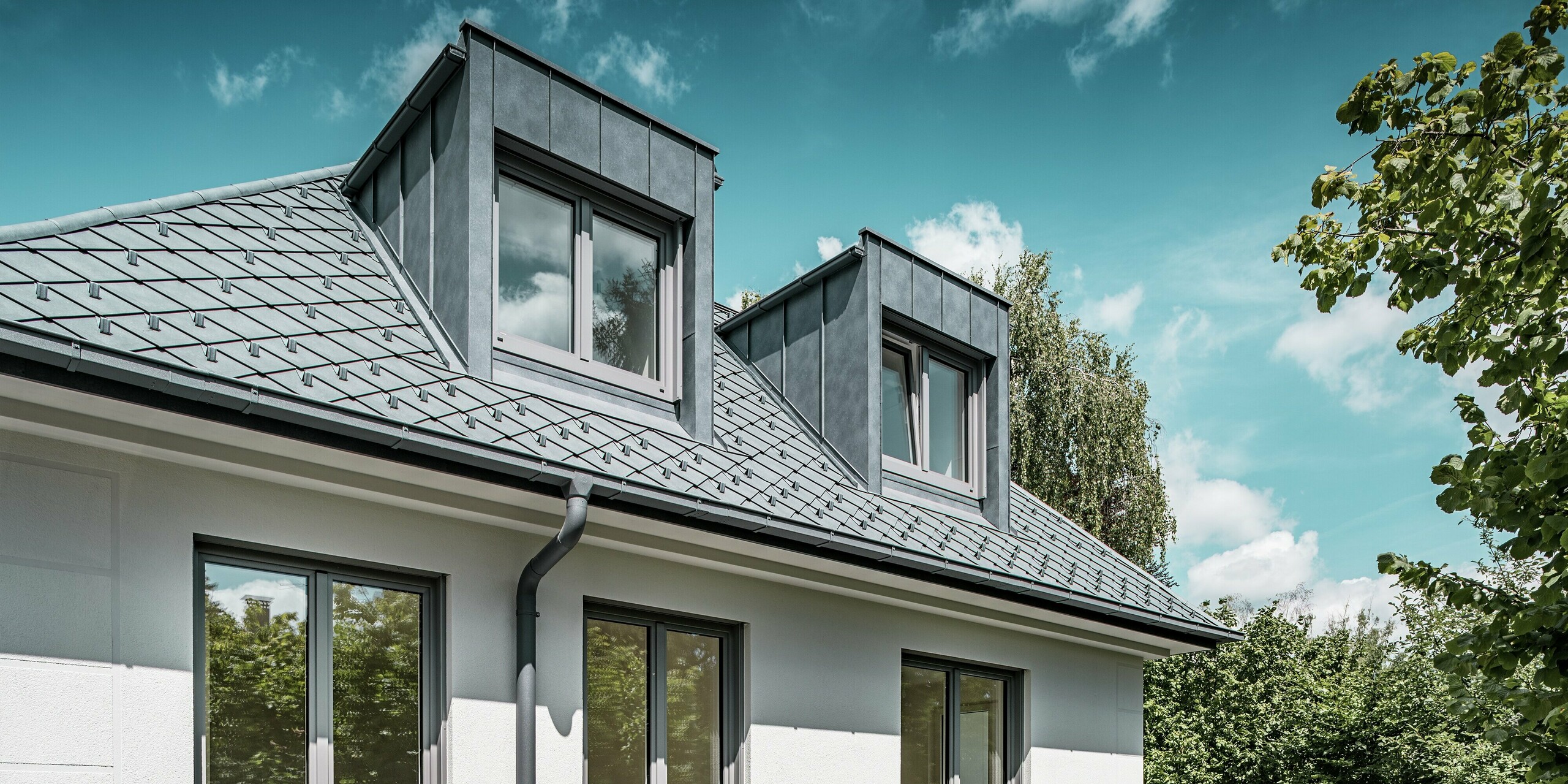 Elegante Perspektive auf ein Mehrfamilienhaus mit markanten Flachdachgauben in PREFALZ und einem präzise gedeckten Dach aus PREFA Dachrauten 29 × 29 in der Farbe P.10 Steingrau. Die Dacharchitektur fügt sich harmonisch in das Gesamtbild ein und wird von der modernen, klaren Linienführung und den großen Fensterflächen ergänzt. Die Gauben aus PREFALZ bieten nicht nur funktionalen Raum, sondern betonen durch ihr minimalistisches Design die hochwertige Bauweise. Die langlebigen Aluminiumprodukte garantieren Witterungsbeständigkeit und eine moderne Ästhetik.