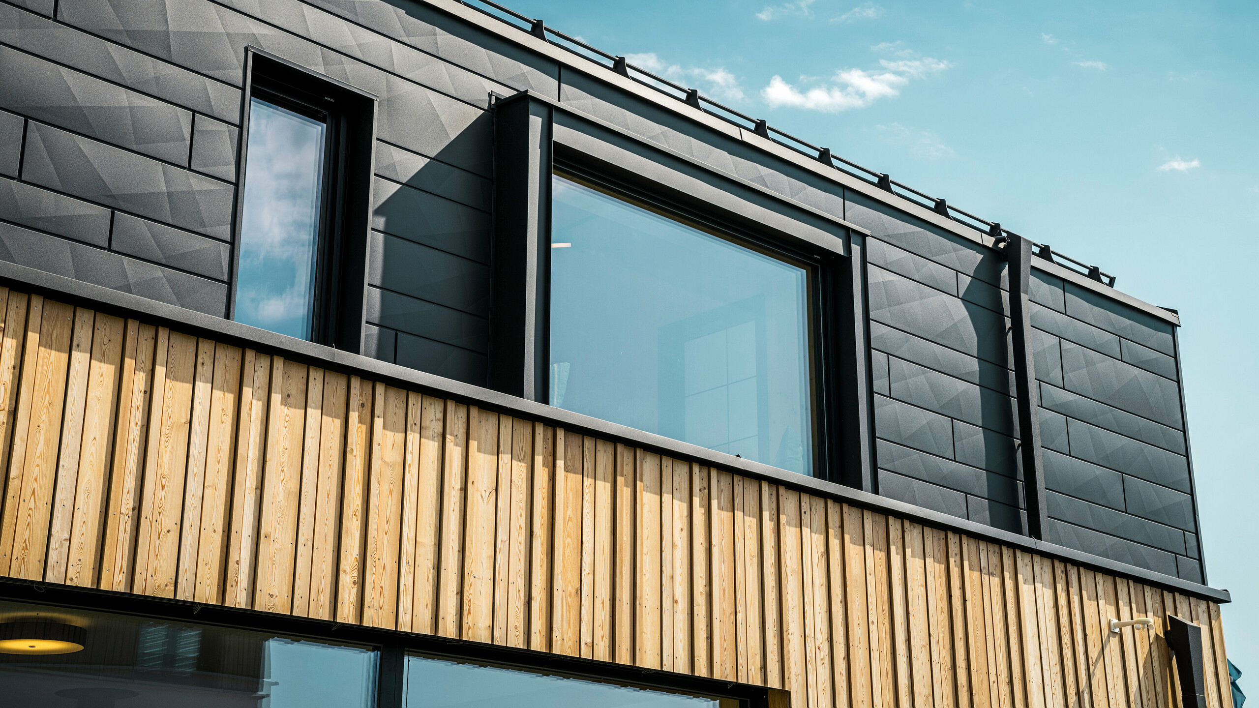 Zbliżenie na górną kondygnację domu modelowego Autark w centrum domów prefabrykowanych Blaue Lagune w Vösendorfie (AT). Elewacja wykonana jest z paneli aluminiowych PREFA Siding.X w kolorze P.10 czarny, które wyróżniają się specjalnym wyglądem z różnymi kątami. Poniżej znajduje się stylowa okładzina drewniana, która stanowi harmonijny kontrast z ciemnym aluminium. Duże okna wpuszczają do wnętrza budynku dużo światła i podkreślają nowoczesny design.