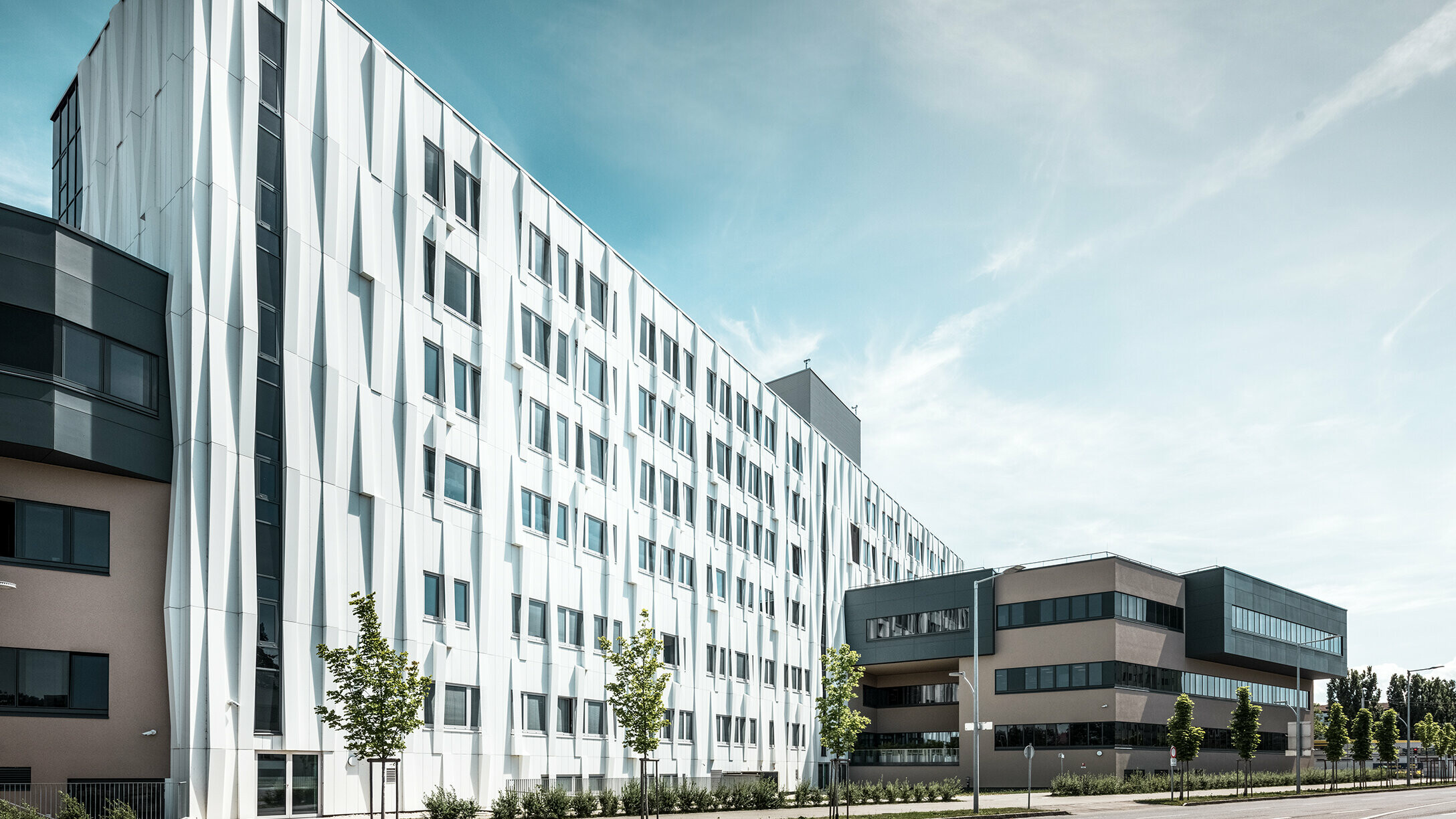 Widok z boku kliniki uniwersyteckiej w St. Pölten w Dolnej Austrii. Solidna aluminiowa fasada sprawia, że budynek przyciąga wzrok w mieście. Na pierwszym planie widać ulicę z kilkoma drzewami.