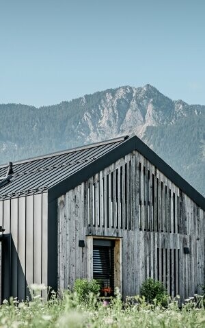 Moderner, funktionaler Wohnbau mit einer Kombination aus Holzfassade und PREFA Band- und Blechmaterial für das Dach. Die klare Architektur wird durch die elegante, metallische Dach- und Fassadenverkleidung unterstrichen, die ideal für anspruchsvolle Projekte ist. Das Bild veranschaulicht die vielseitigen Anwendungsmöglichkeiten der PREFA Produkte im Bereich Bänder und Bleche.
