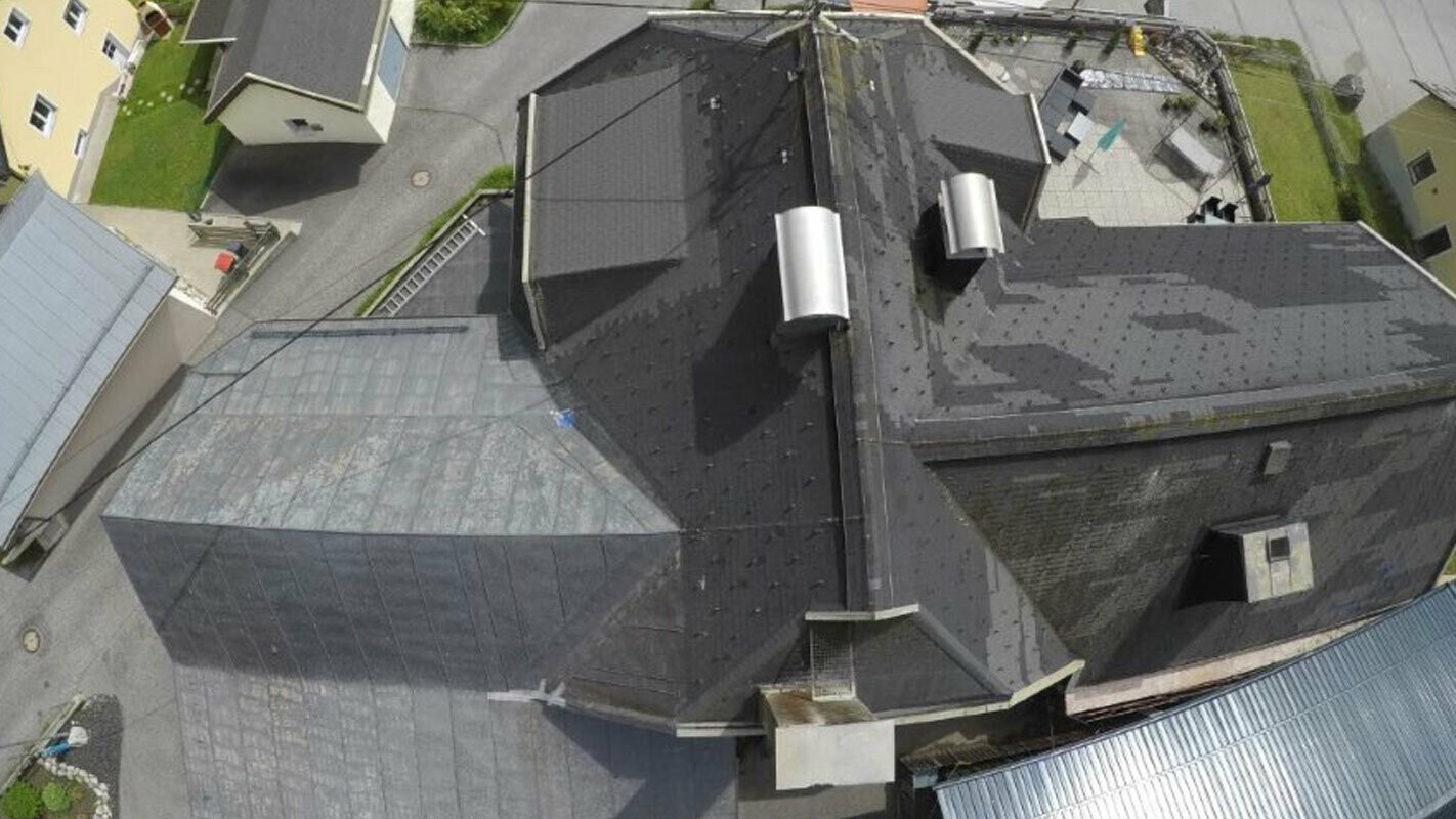 Perspektywa z lotu ptaka starego dachu z wieloma szczegółami, koszami dachowymi, oknami dachowymi i kominami. Stare pokrycie dachu było wielokrotnie modernizowane i jest plamiste.