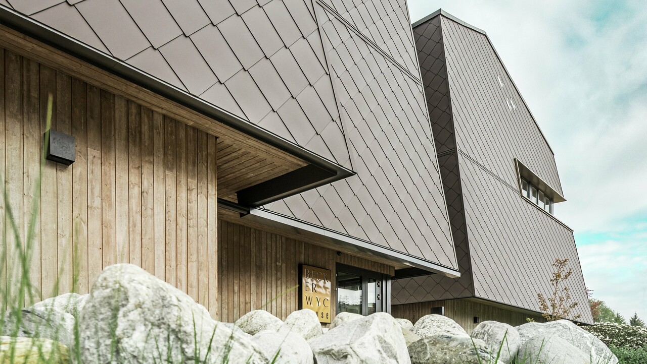 Widok z boku hotelu, fasada parteru jest pokryta pionowo drewnem w ciemnym kolorze. Powyżej znajduje się występ elewacji, od którego fasada jest pokryta aluminiowymi panelami w kolorze orzechowego brązu.