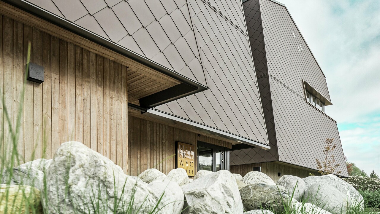 Widok z boku hotelu, fasada parteru jest pokryta pionowo drewnem w ciemnym kolorze. Powyżej znajduje się występ elewacji, od którego fasada jest pokryta aluminiowymi panelami w kolorze orzechowego brązu.