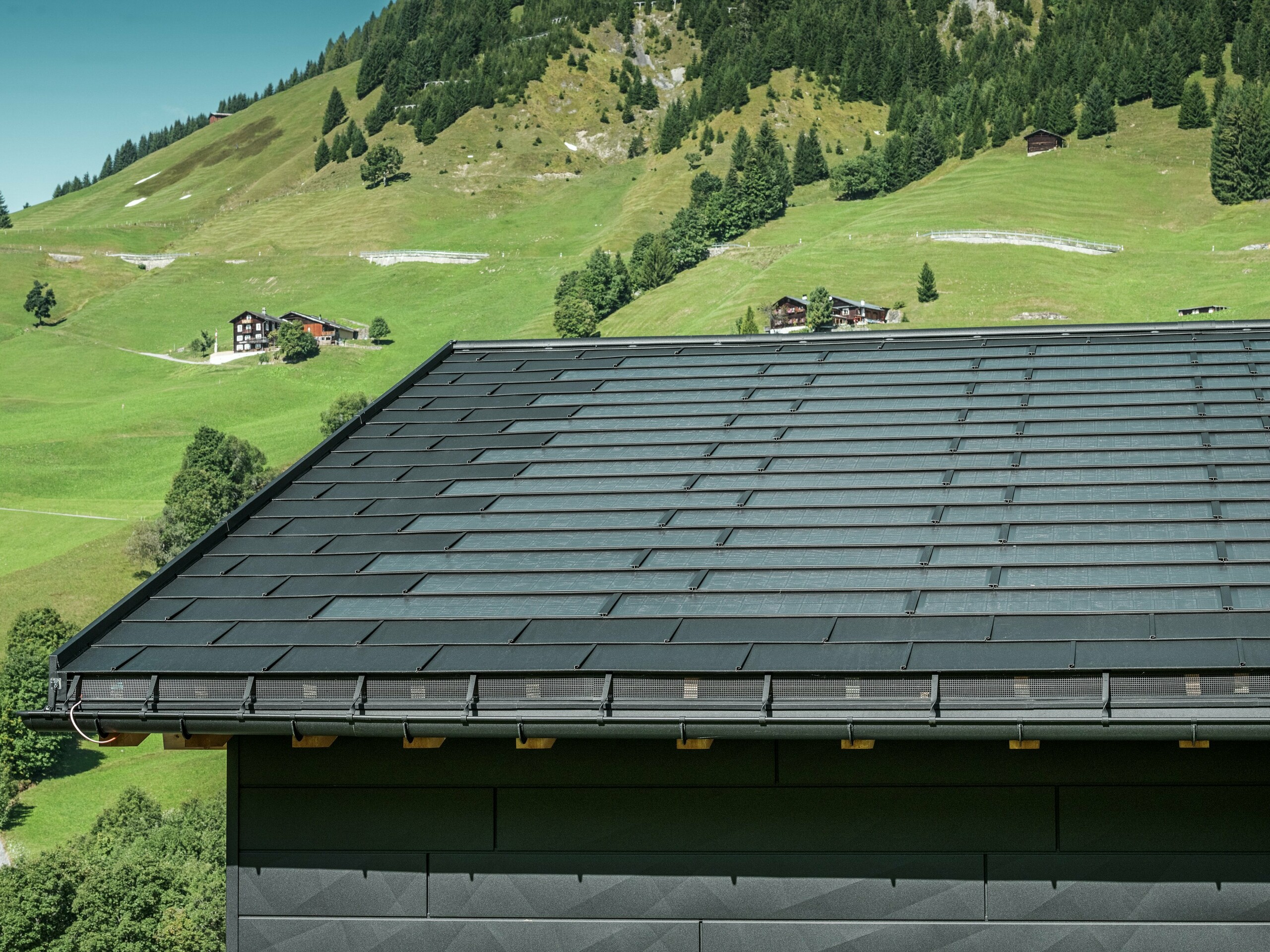 Zbliżenie na okap nowoczesnego domu jednorodzinnego w alpejskiej okolicy Ascharina w Szwajcarii. Dach pokryto innowacyjną dachówką solarną PREFA oraz tradycyjną dachówką klasyczną R.16, obie w kolorze P.10 czarny. Niezawodne odwodnienie dachu zapewniają rynna półokrągła PREFA oraz rura spustowa PREFA. Na dolnej krawędzi zdjęcia widoczne są charakterystyczne panele elewacyjne PREFA Siding.X. Wyraziste linie produktów aluminiowych podkreślają nowoczesny design w malowniczym górskim krajobrazie.
