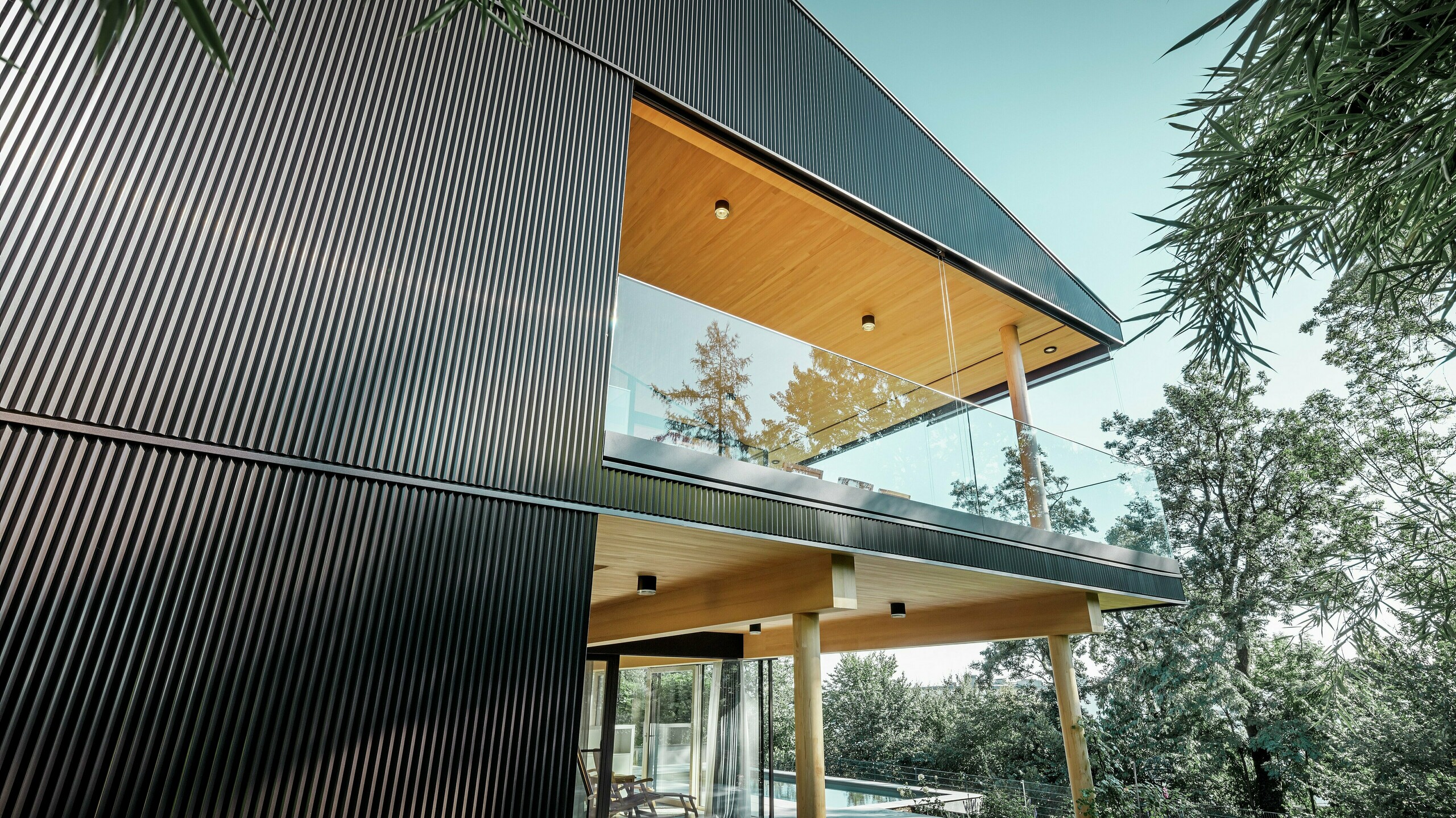 Widok z boku domu jednorodzinnego, fasada pokryta ciemnym Profilem trójkątnym PREFA. Widoczny jest taras i przestronny balkon. Sufit lub podbitka zostały zaprojektowane z drewnianych paneli.