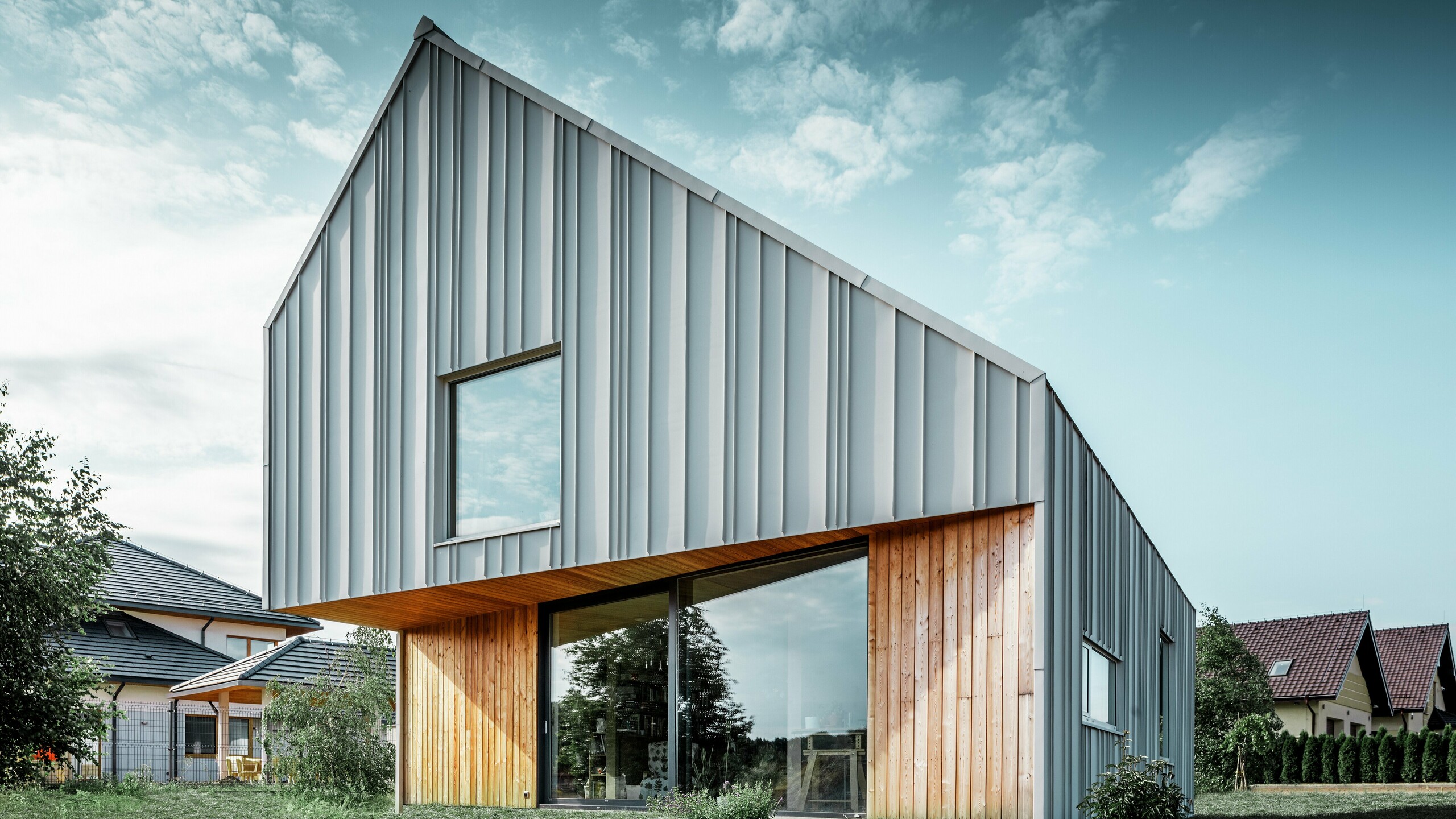 Nowoczesny dom jednorodzinny z dwuspadowym dachem bez okapów i aluminiową fasadą PREFA w kolorze białym. Elewacja drewniana jest połączona z pionowymi panelami drewnianymi na parterze. 