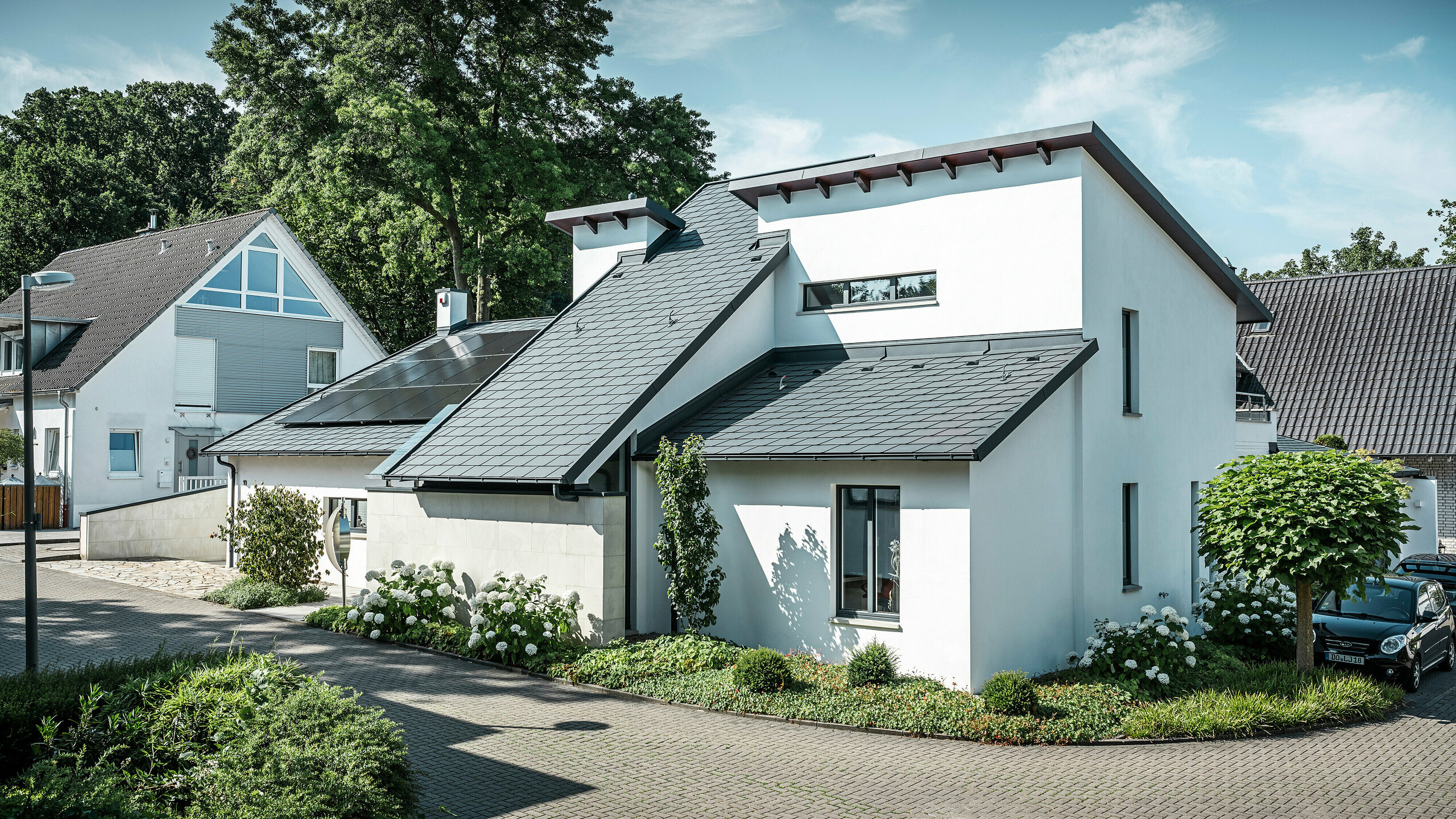 Nowoczesny dom jednorodzinny z białą elewacją pokrytą tynkiem i ciemnym dachem skośnym PREFA z aluminium. Na dachu widoczne są zintegrowane moduły fotowoltaiczne i aluminiowe odwodnienie dachu w kolorze antracyt. Budynek położony jest na zadbanym osiedlu mieszkaniowym z zadbanymi ogrodami przed domami, brukowanym podjazdem i otaczającymi domami jednorodzinnymi.