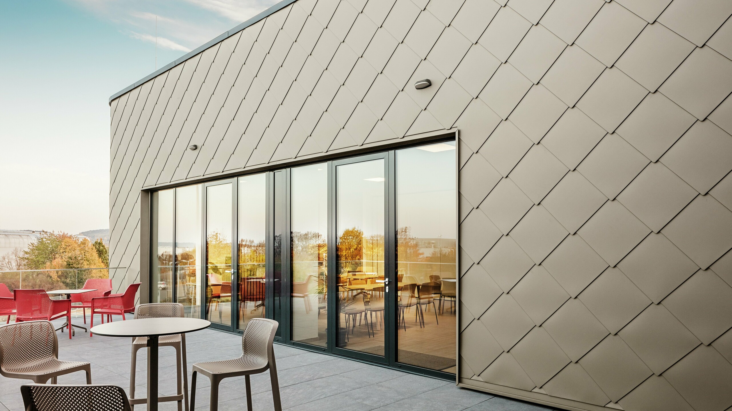 [Translate to Polski:] Die Fassade des neuen PREFA Standortes in Törökbálint besticht durch die eleganten PREFA Wandrauten 44 × 44 in Bronze. Die präzise verlegte Aluminiumfassade sorgt für ein modernes, dynamisches Erscheinungsbild und bietet gleichzeitig maximalen Schutz vor Witterungseinflüssen. Die großzügige Glasfront verbindet Innen- und Außenbereich und schafft auf der großzügigen Terrasse eine helle und einladende Atmosphäre.