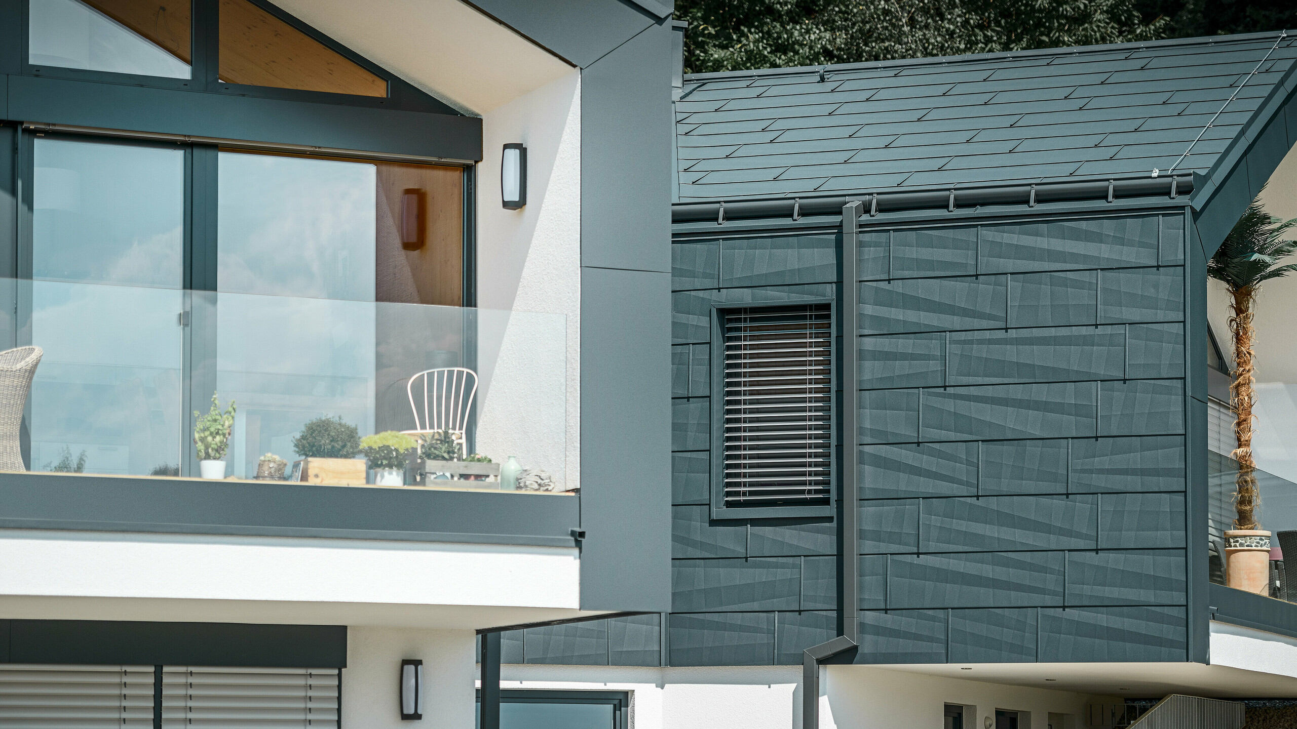 Zdjęcie przedstawia nowoczesny dom bliźniaczy z elegancką elewacją z aluminium. Elewacja utrzymana jest w ciemnym odcieniu antracytu i ma teksturowaną powierzchnię dzięki panelom dachowym i panelom elewacyjnym PREFA FX.12. Na dachu zamontowano rynnę leżącą i rurę kwadratową jako rura spustowa PREFA. Na piętrze widoczny jest balkon z balustradą szklaną, natomiast na parterze znajdują się okna z żaluzjami lamelowymi. Architektura budynku jest przejrzysta i minimalistyczna, a w tle widać drzewa, które tworzą naturalną atmosferę.