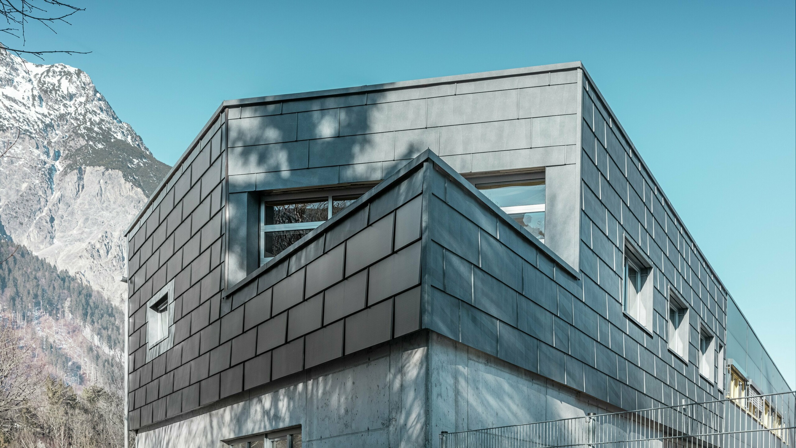 Budynek firmy Spenglerei RÜMA w Vorarlbergu zachwyca nowoczesną elewacją wykonaną z dachówki solarnej PREFA i betonu licowego. Otoczony górami i drzewami, których cienie odbijają się na ścianie, stoi pod czystym, błękitnym niebem. Ostry narożnik budynku i panel elewacyjny FX.12 dodatkowo podkreślają jego charakterystyczny wygląd.