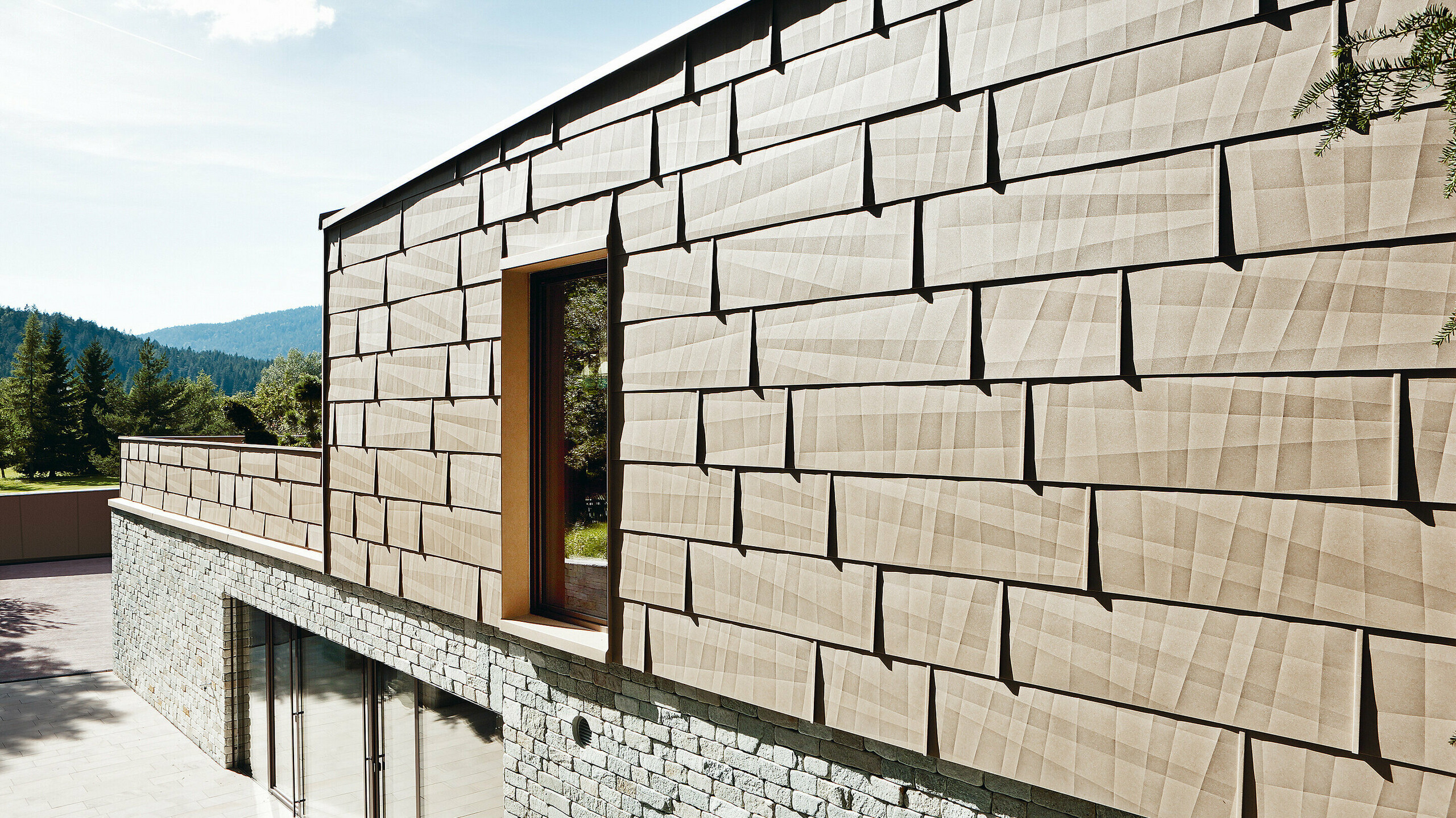 Ta fasada zewnętrzna stanowi połączenie paneli elewacyjnych PREFA FX.12 w kolorze P.10 brąz piaskowy z kamienną fasadą piwnicy.