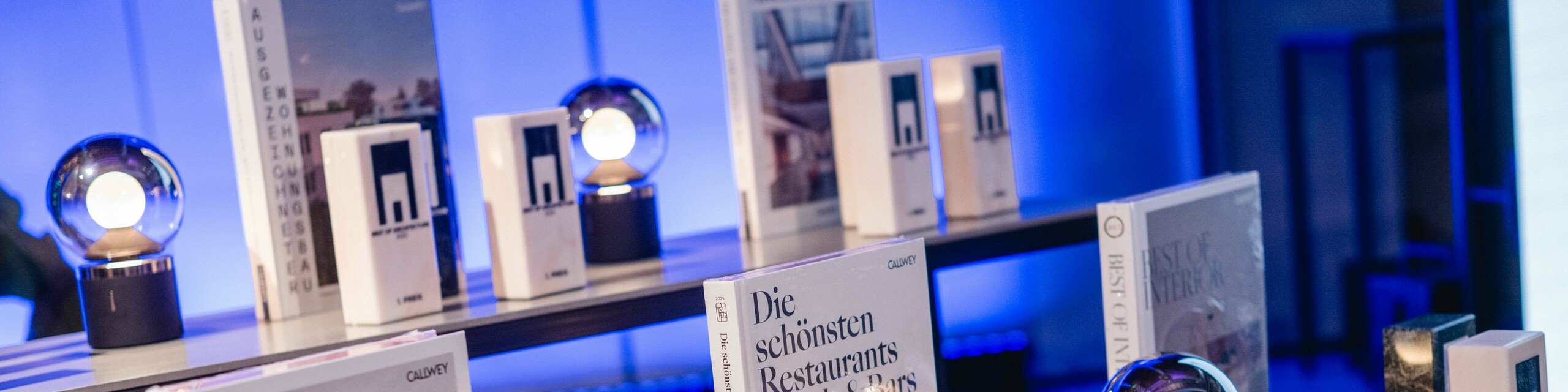 Wystawa nagród z konkursu „Budynek mieszkalny roku” 2025: kilka eleganckich trofeów z logo „Best of Architecture” jest wyeksponowanych na nowoczesnym stole, obok wysokiej jakości książek o architekturze i designie, takich jak „Best Workspaces” i „Die schönsten Restaurants Hotels & Bars” (Najpiękniejsze restauracje, hotele i bary). Scena jest skąpana w nastrojowym niebieskim świetle, które oddaje ekskluzywność, innowacyjność i doskonałość architektoniczną.