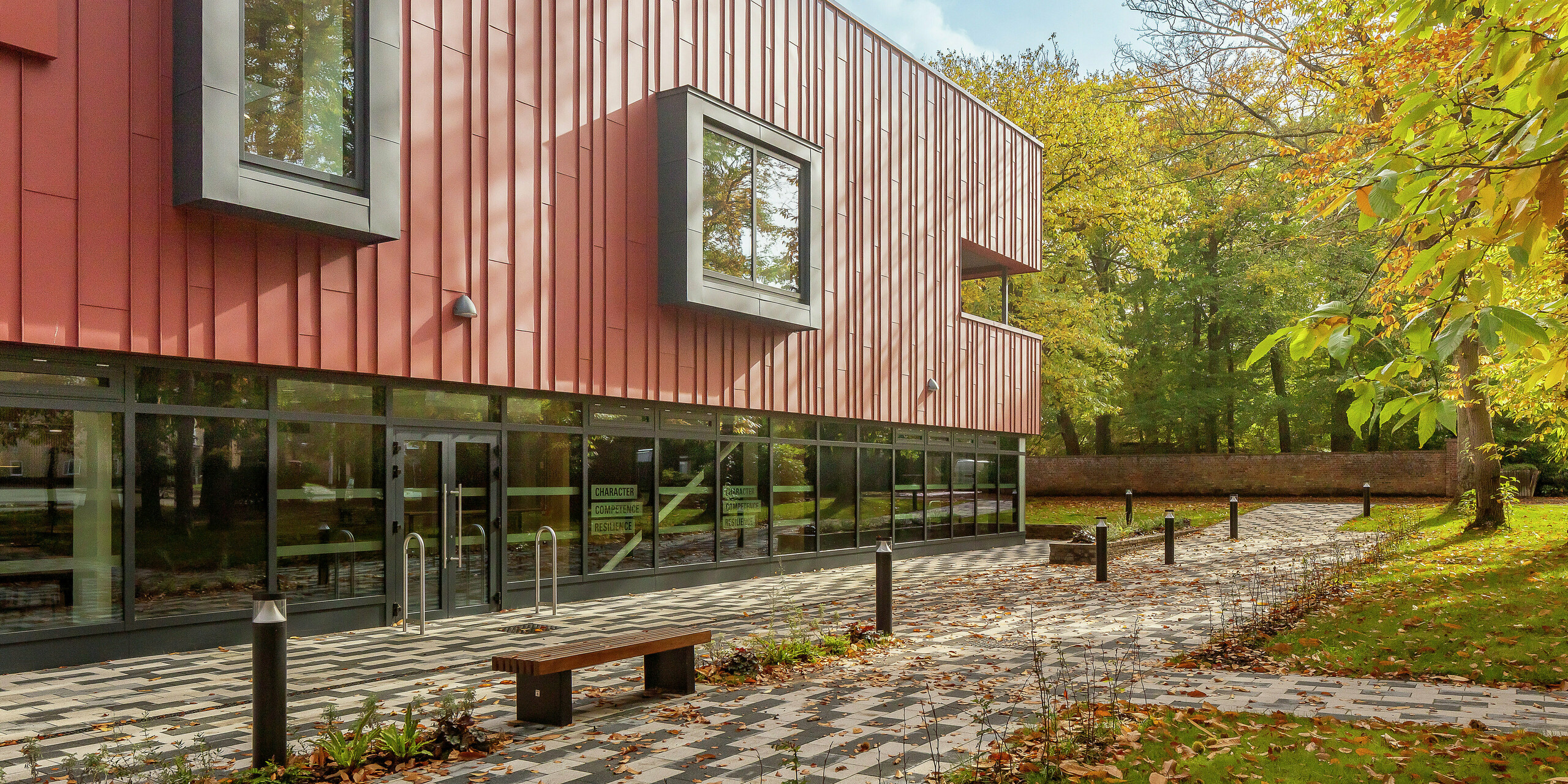 Harmonische Architektur im Grünen: Die moderne Stehfalzfassade des Soldiers' Centre in Pirbright besteht aus PREFALZ in P.10 Oxydrot. Die robuste Aluminiumverkleidung fügt sich perfekt in die herbstliche Umgebung ein. Auffällige, auskragende Fensterrahmen in P.10 Anthrazit setzen stilvolle Akzente. Große Glasflächen im Erdgeschoss sorgen für Transparenz und Offenheit. Der gepflasterte Vorplatz mit Sitzbank lädt zum Verweilen ein - eine gelungene Verbindung von Design, Funktionalität und Natur.