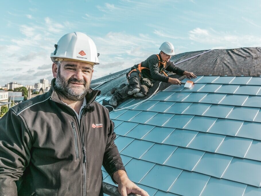 Zwei Handwerker auf einem Dach, der eine posiert mit einem Lächeln, während der andere an der Installation der PREFA Dachschindeln arbeitet. Beide tragen Schutzhelme und Sicherheitskleidung. Das Bild illustriert einen Erfahrungsbericht von Verlegepartnern, die ihre Expertise und die Zusammenarbeit mit PREFA bei der Montage von Dachsystemen teilen. Es spiegelt die Professionalität und den Teamgeist wider, die für eine erfolgreiche Umsetzung von Projekten notwendig sind.