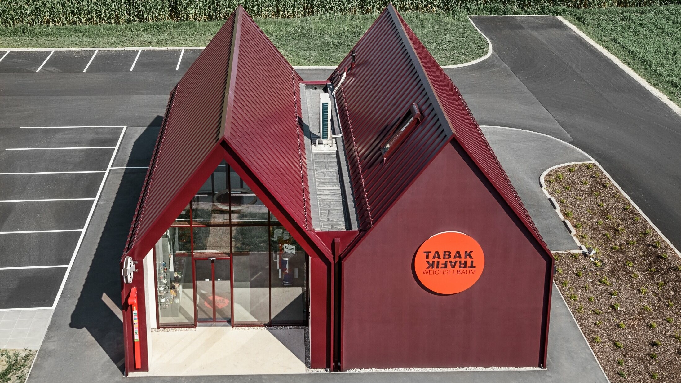 Nowoczesny sklep tytoniowy z dachem dwuspadowym bez okapu, z okładziną aluminiową PREFA wykonaną w systemie pionowych i kątowych zakładek w kolorze specjalnym czerwono-czarnym