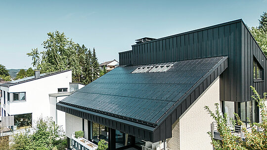 Nowoczesna architektura domu jednorodzinnego w Meschede w Niemczech, z dachem aluminiowym PREFALZ w kolorze P.10 czarny i zintegrowanymi modułami fotowoltaicznymi szkło/szkło. Dach z przesunięciem łączy w sobie ponadczasowy design z zrównoważoną technologią fotowoltaiczną. Innowacyjne moduły solarne PREFALZ zapewniają moc 18 kWp. Bezramowe moduły oraz eleganckie aluminium pokrycie dachowe i elewacyjne tworzą estetyczną i funkcjonalną całość, która harmonijnie wtapia się w zielone otoczenie.